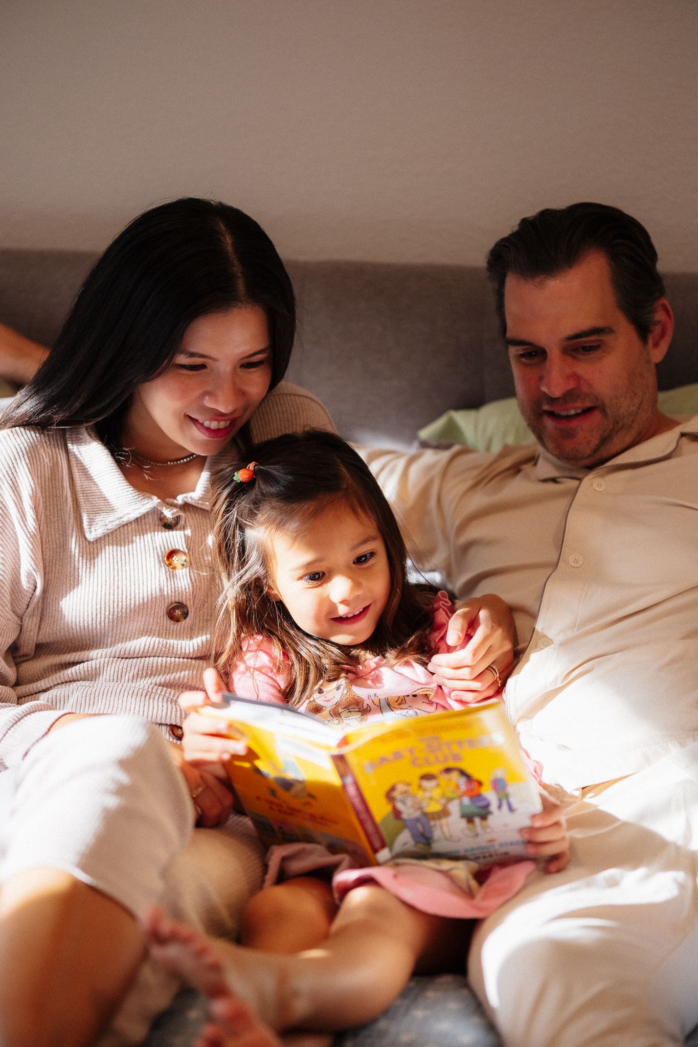 A family enjoys reading a children's book together while relaxing on a bed in warm lighting.