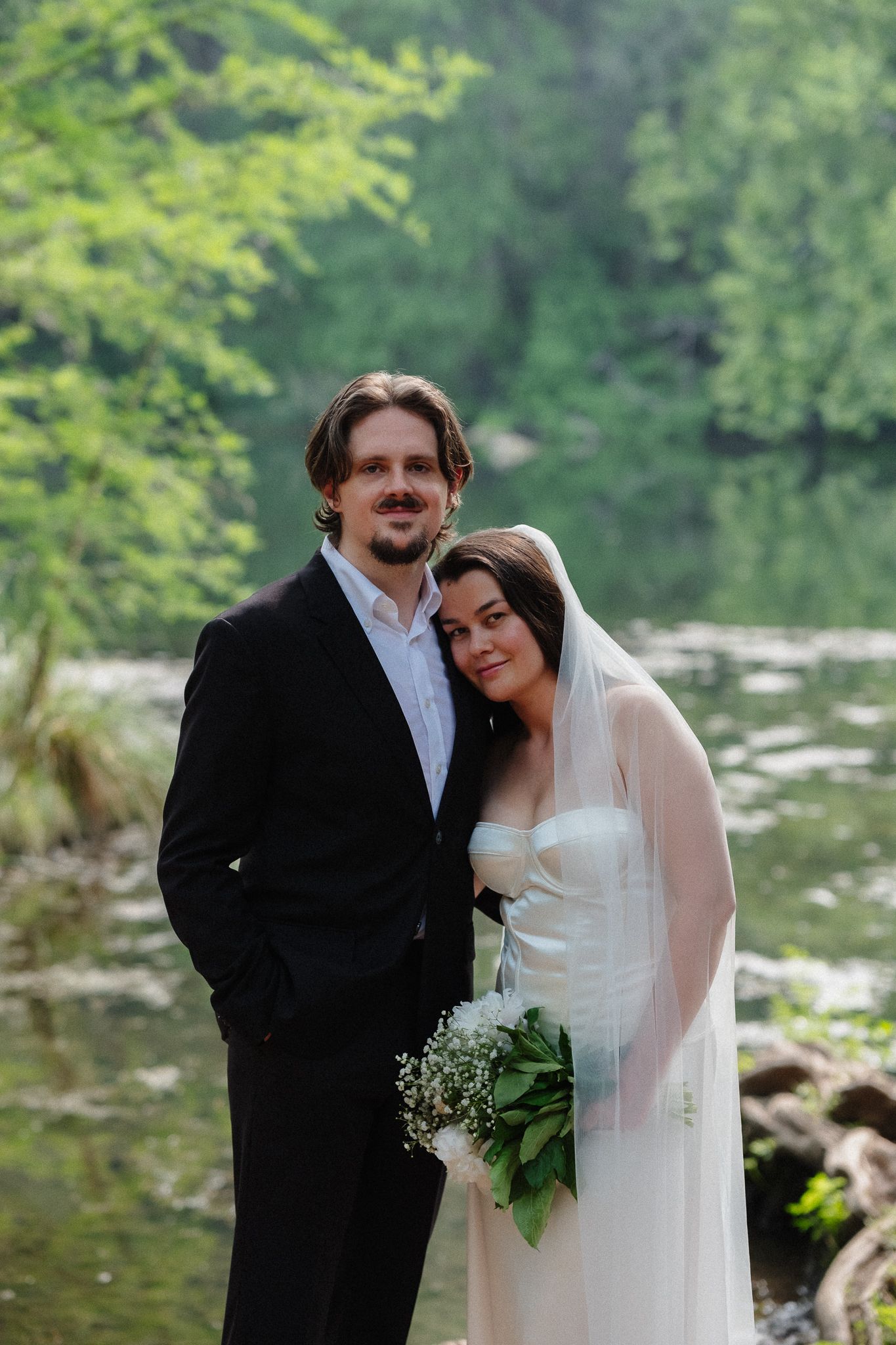 Bride and groom posing outdoors near a serene river, surrounded by lush green trees, bride holding a bouquet.