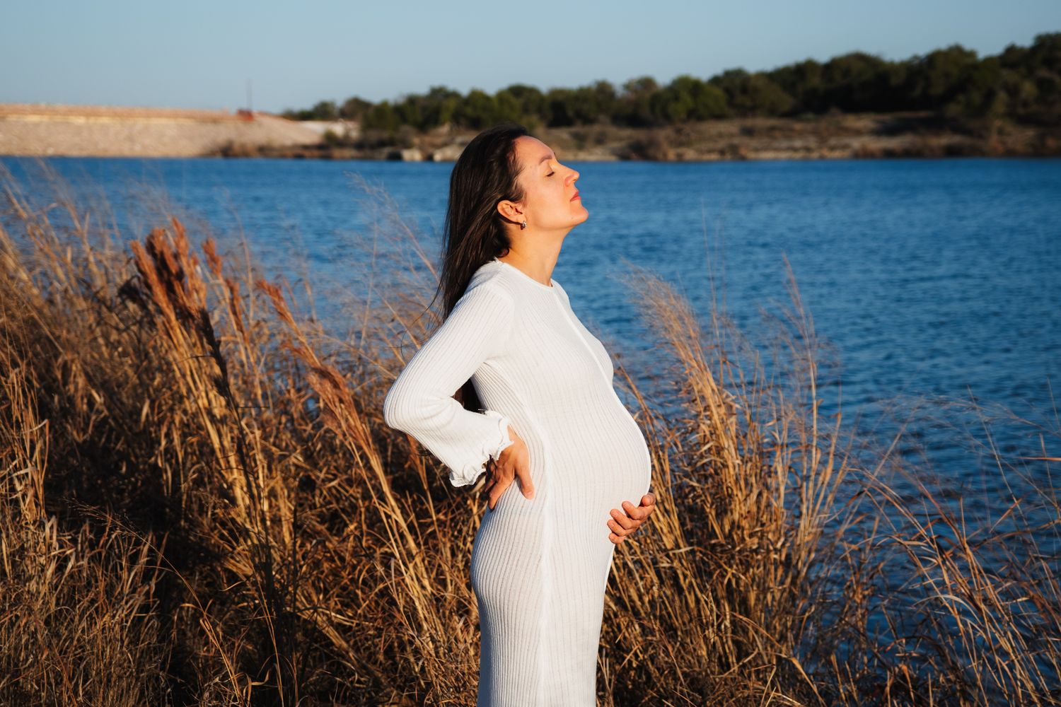 Silhouetted figure in white dress stands by lakeside with tall grasses during maternity shoot at golden hour.