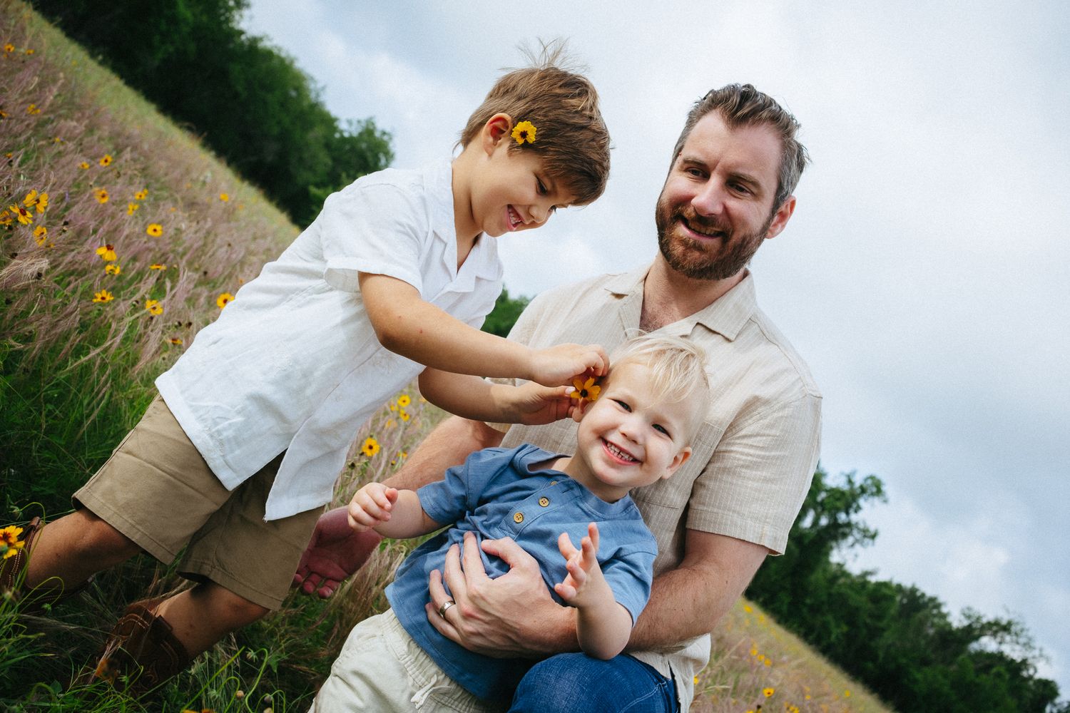 A smiling father holds his toddler while an older child plays with flowers in a sunny meadow field.