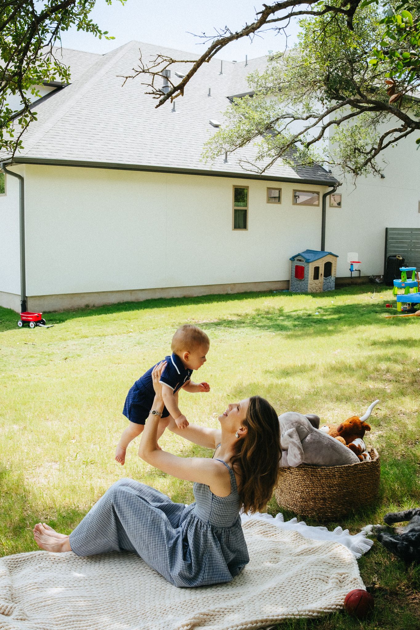 Mother lifting baby in backyard on picnic blanket near house with toys on sunny day.