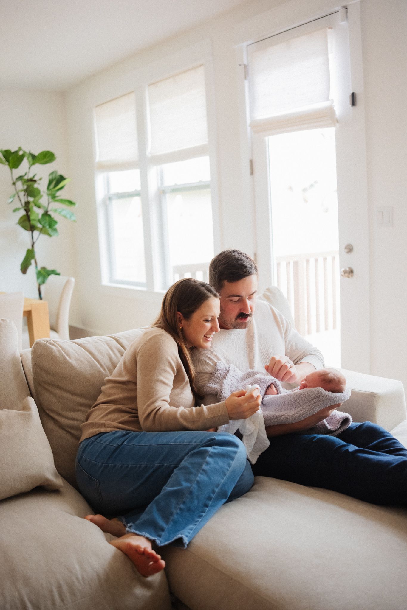 A couple sits on a cozy sofa, lovingly gazing at their newborn baby in a bright, modern living room.
