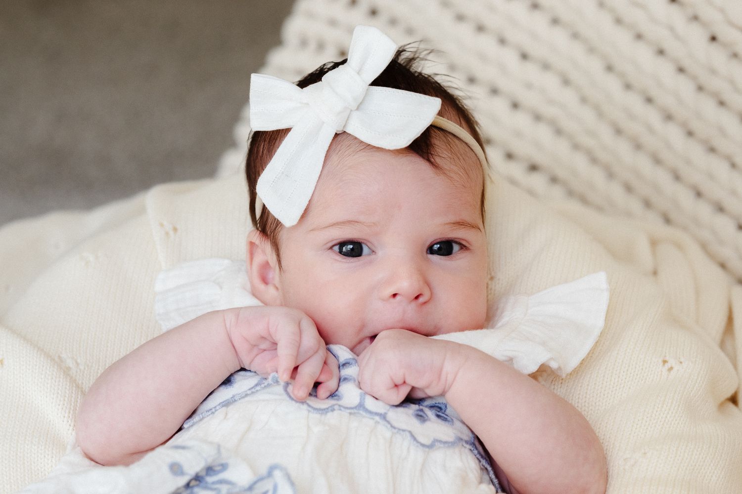 Adorable baby girl with white bow headband lying on cream blanket, chubby hands near mouth, big blue eyes.