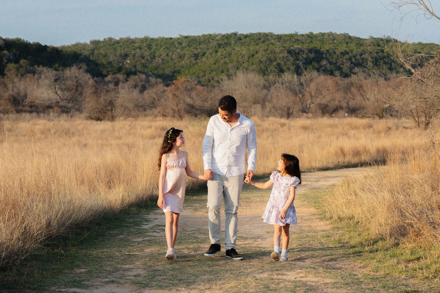 A father walks hand-in-hand with two young daughters on a scenic dirt path through golden fields.
