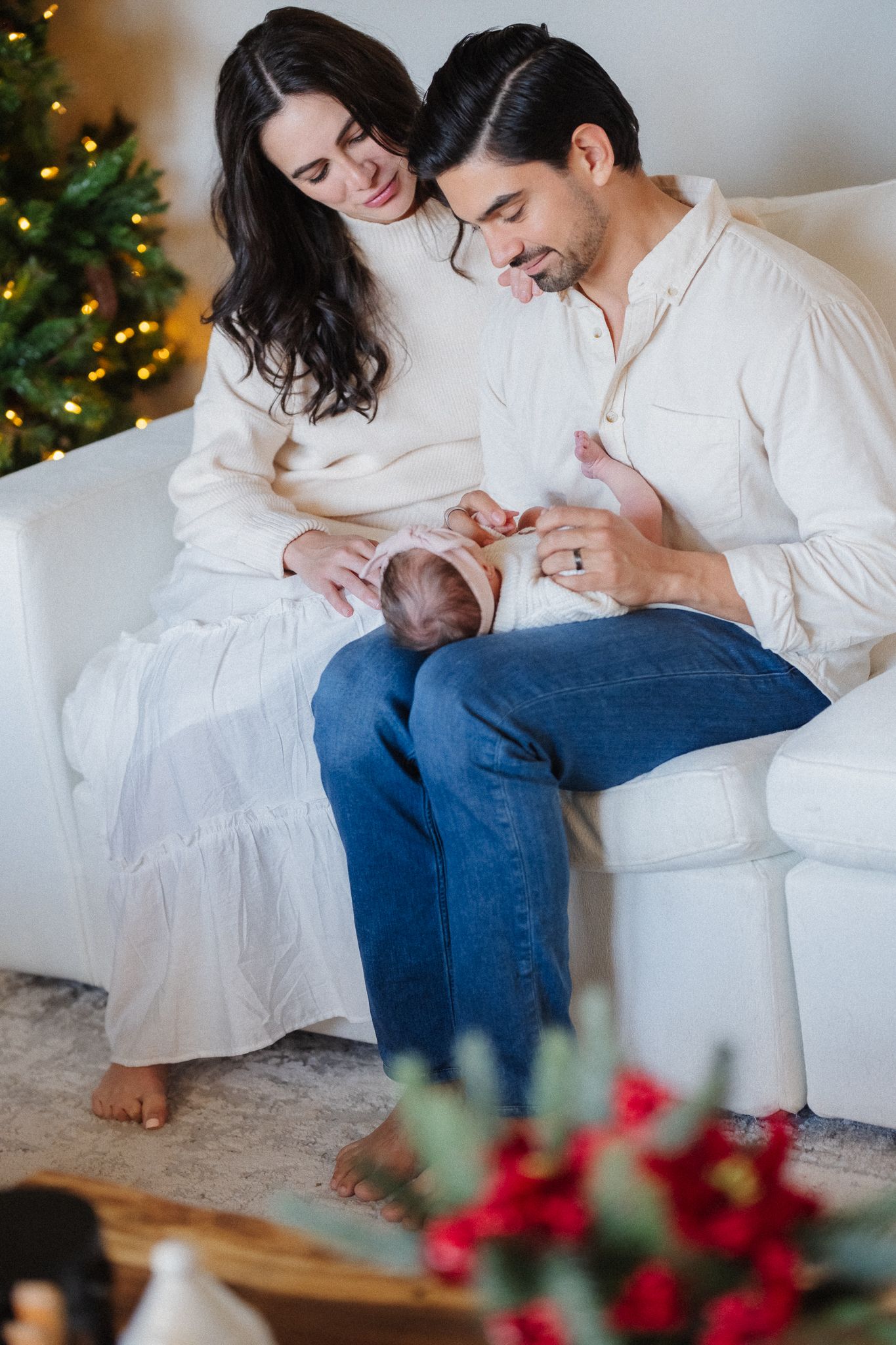 A couple in white outfits sits on a sofa holding their newborn baby near a decorated Christmas tree.