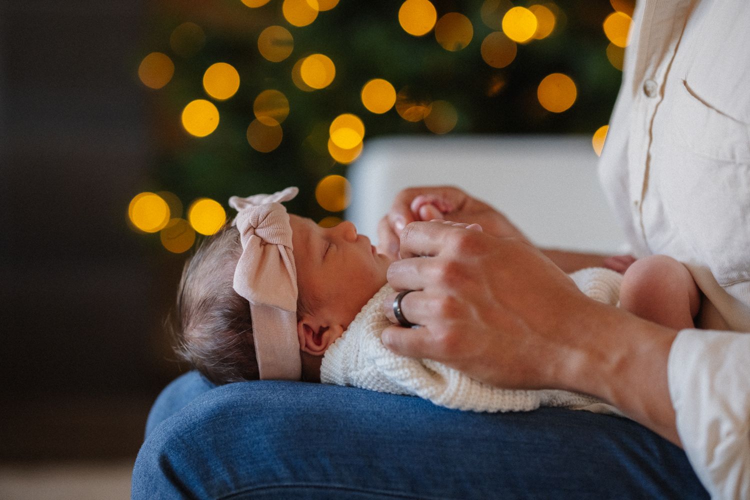 Newborn baby in white knit swaddle with pink bow headband cradled in mothers lap near Christmas tree lights.
