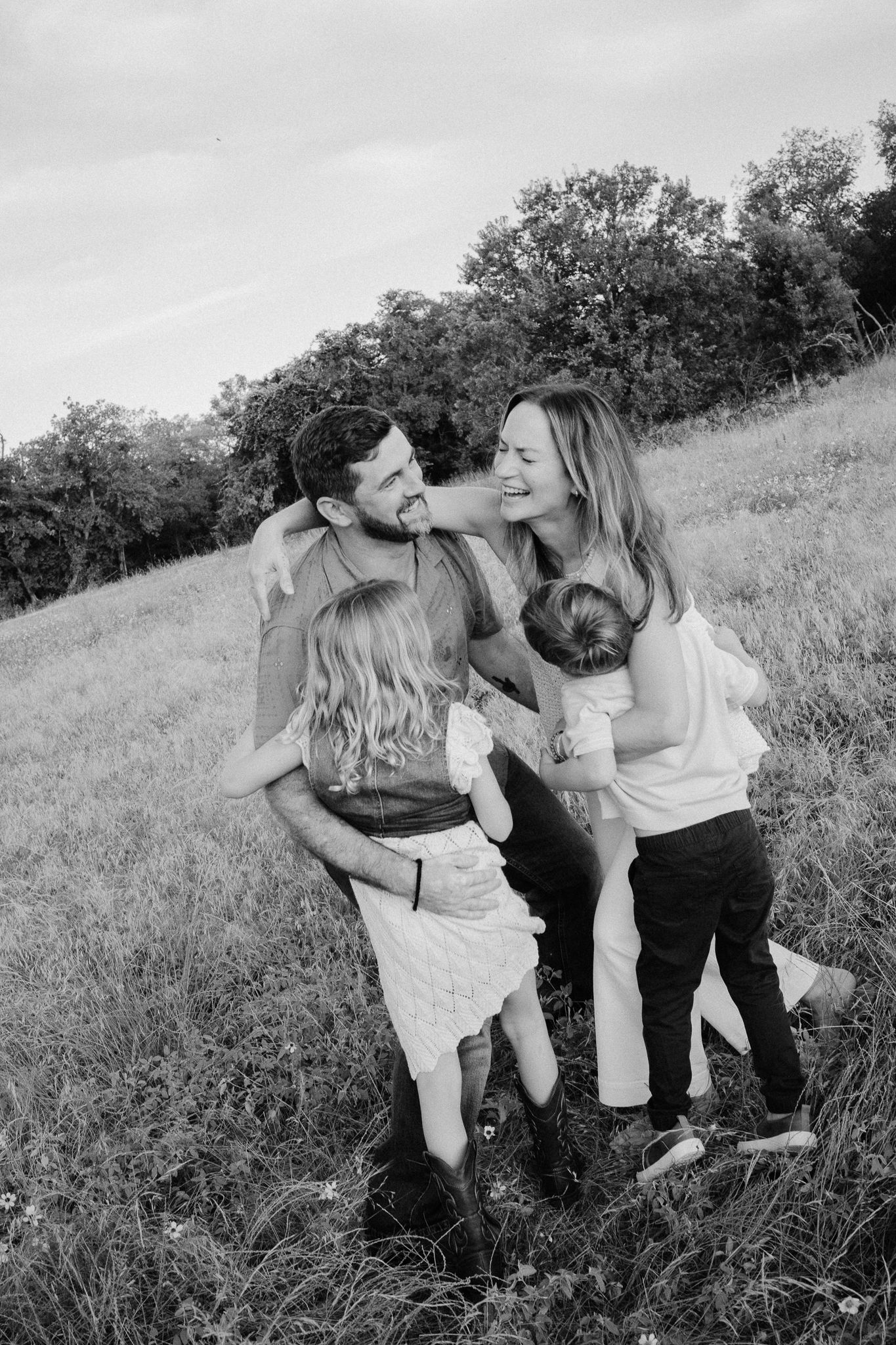 Black and white photo of a happy family of four laughing together in a grassy field with trees in the background.