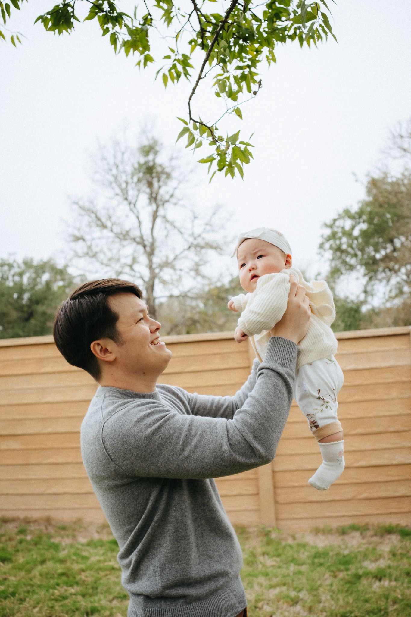 A person in a gray sweater lifts a baby wearing white in a backyard with trees and a wooden fence.