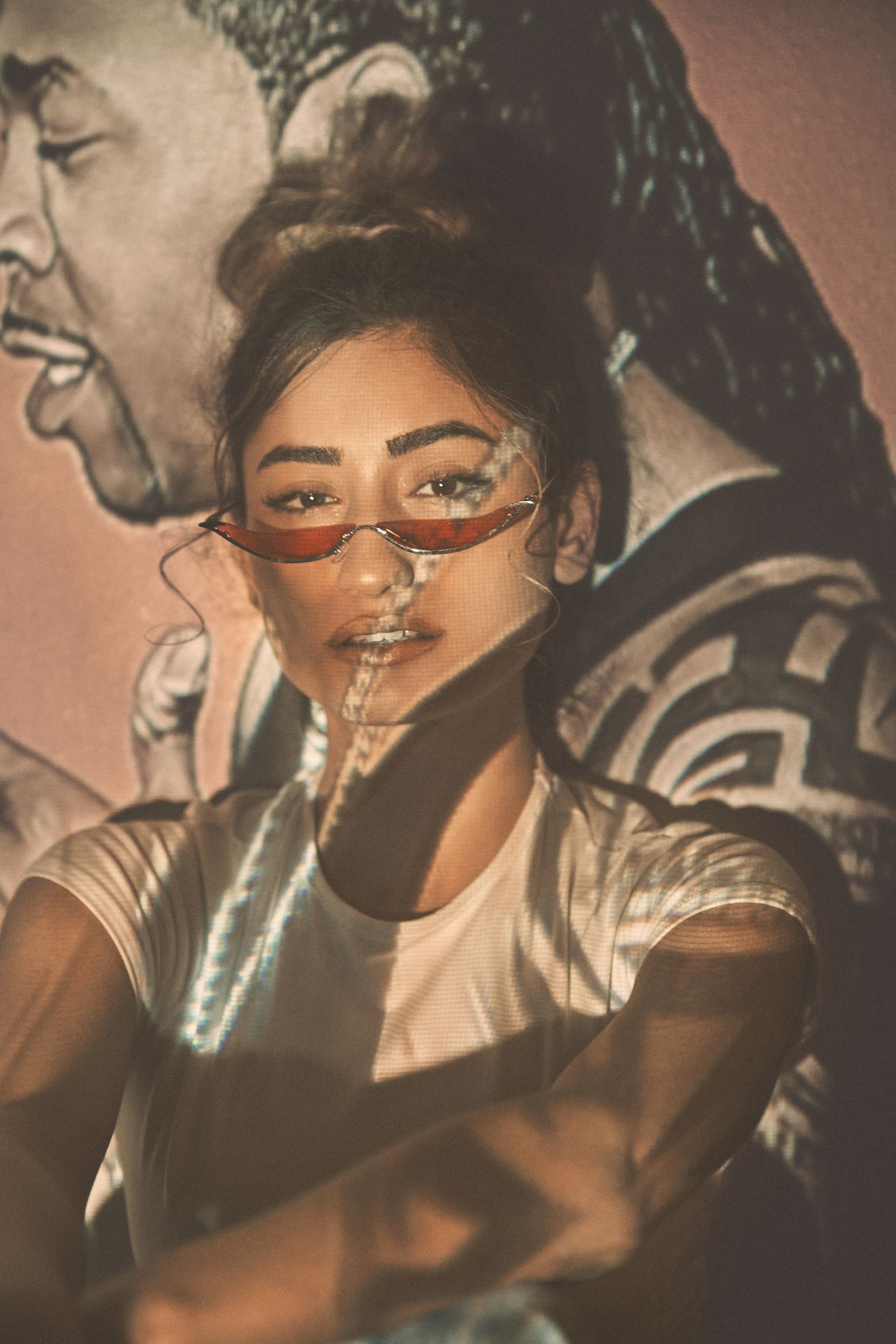 A stylish portrait shot of a person wearing sunglasses and a white t-shirt against a background showing an illustrated mural.