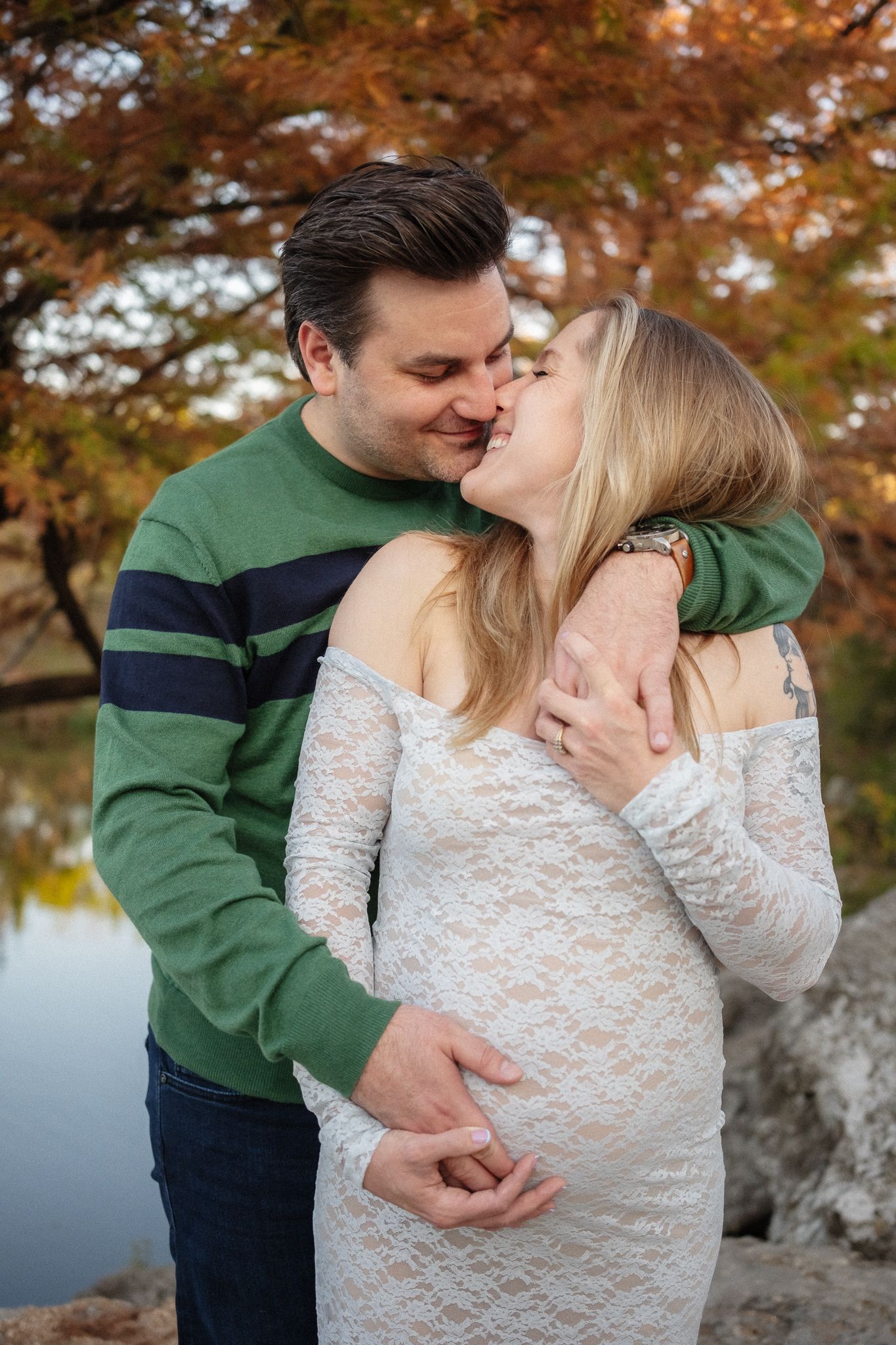 A couple shares an intimate moment outdoors during a maternity photoshoot with autumn foliage and water in the background.