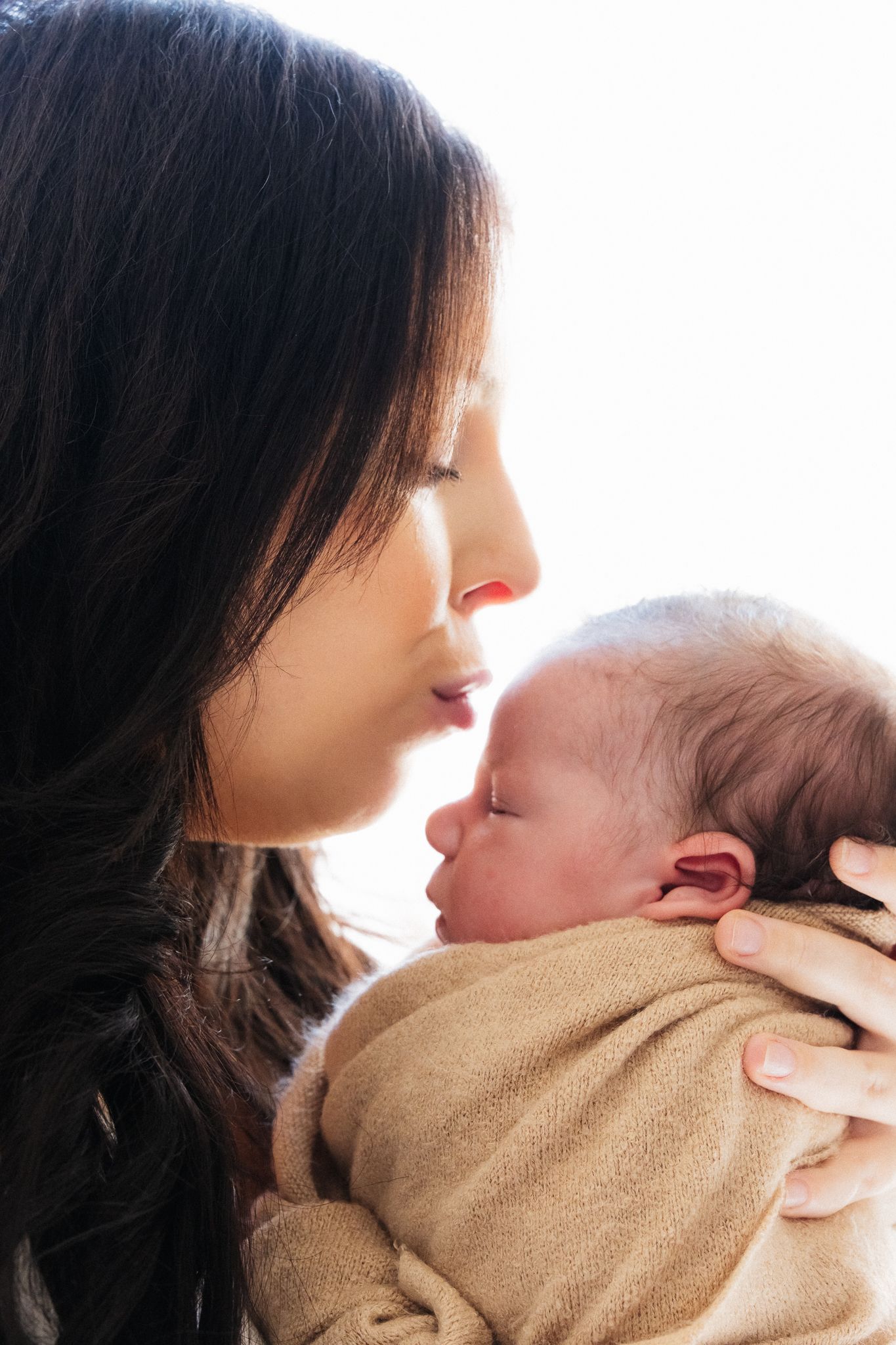A tender moment shows a mother cradling and kissing her newborn baby wrapped in a soft brown blanket.