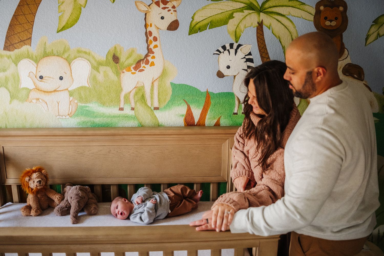 Parents admiring newborn baby in jungle-themed nursery crib with safari animal wall mural and stuffed toys.