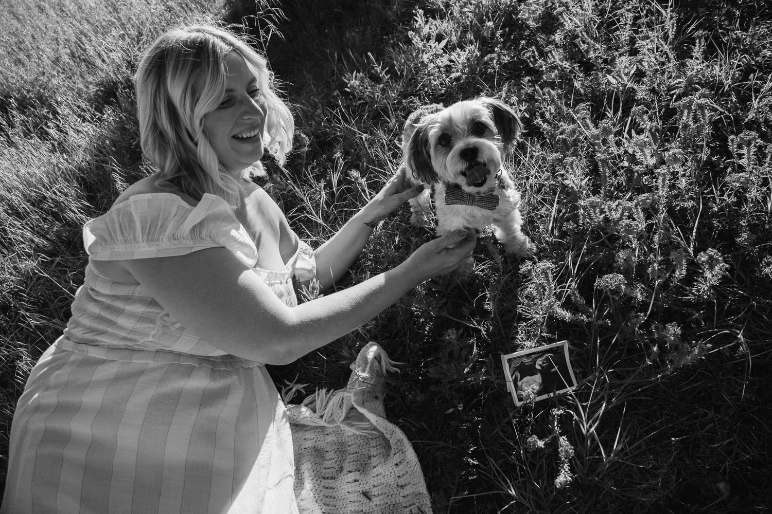 Black and white photo of a pregnant woman in a white dress joyfully holding up a small scruffy dog in an outdoor garden setting.