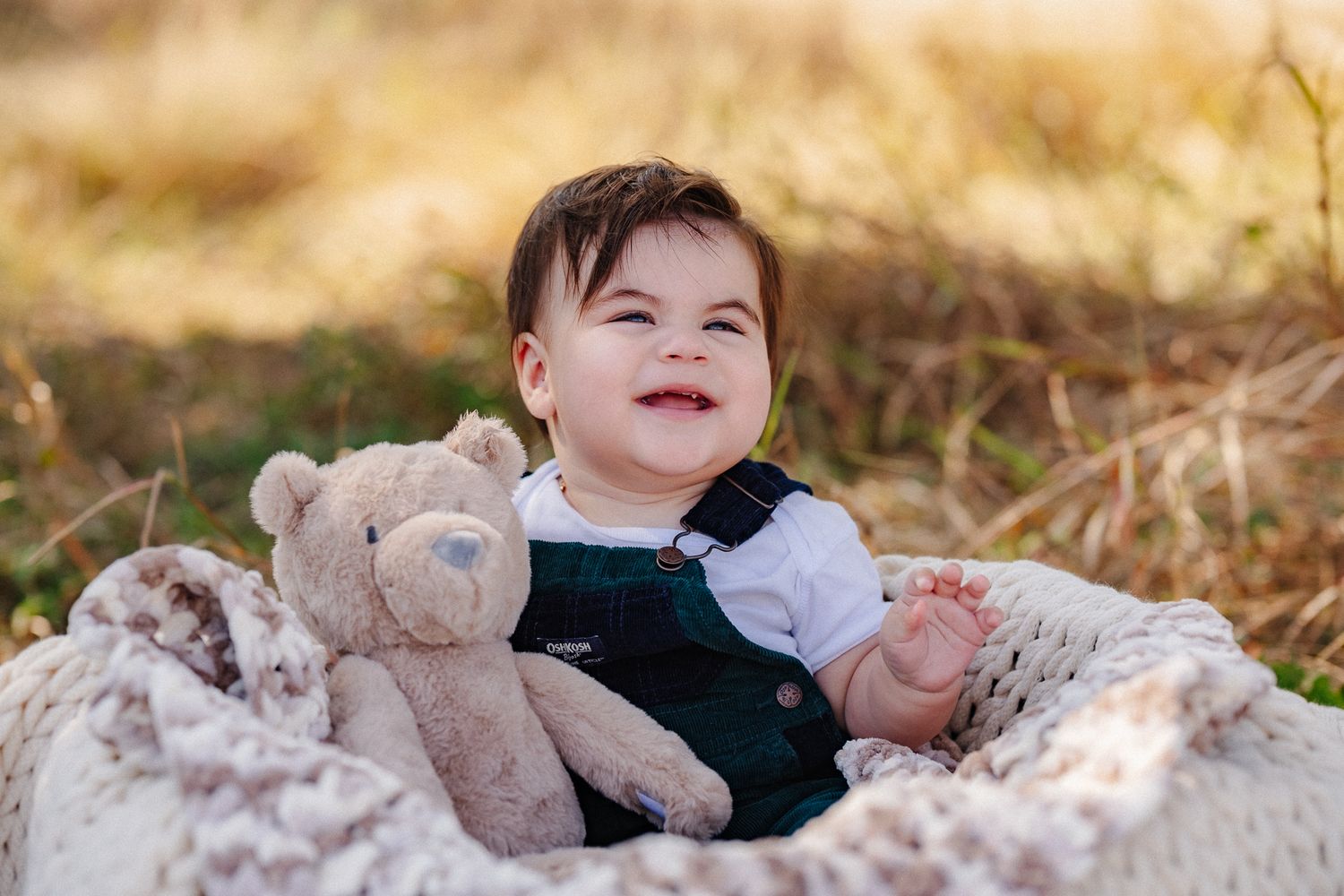 A child lies in a field of golden grass while holding a plush teddy bear during an outdoor photo session.