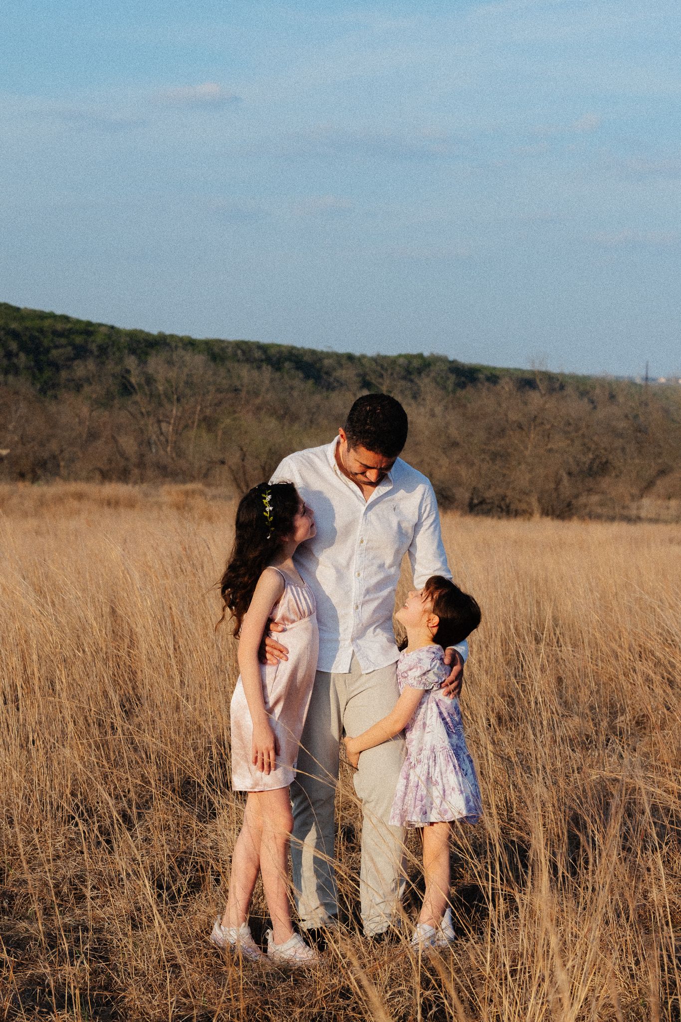 A man stands with two young girls in a golden field, sharing a warm family moment under a blue sky.