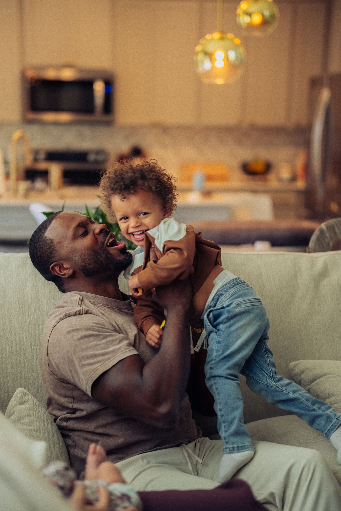 A joyful father lifts his laughing toddler on a cozy sofa in a warm, modern home setting.