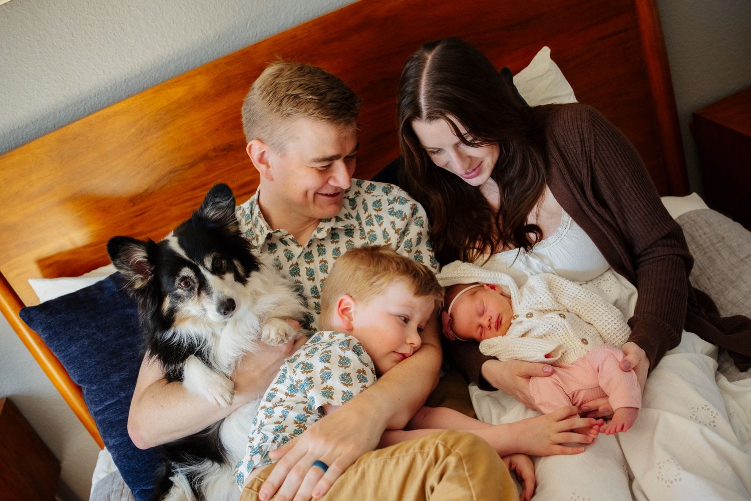 Family with newborn baby, young boy, and border collie dog resting together on a bed in a warm home setting.