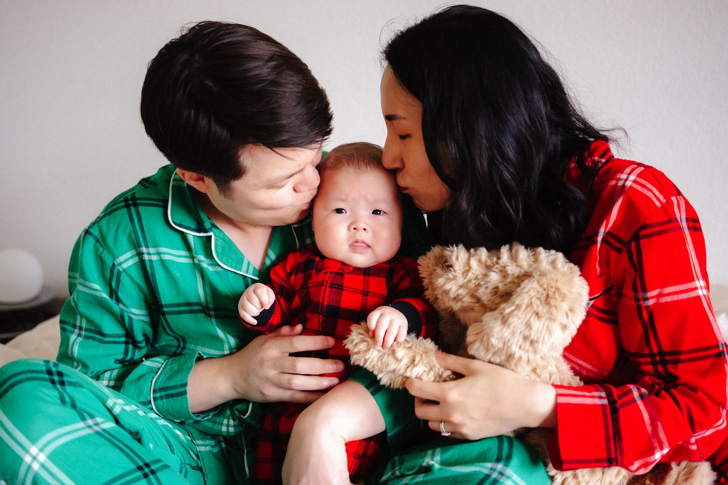 Two parents in matching plaid Christmas pajamas kissing their baby while holding a teddy bear.