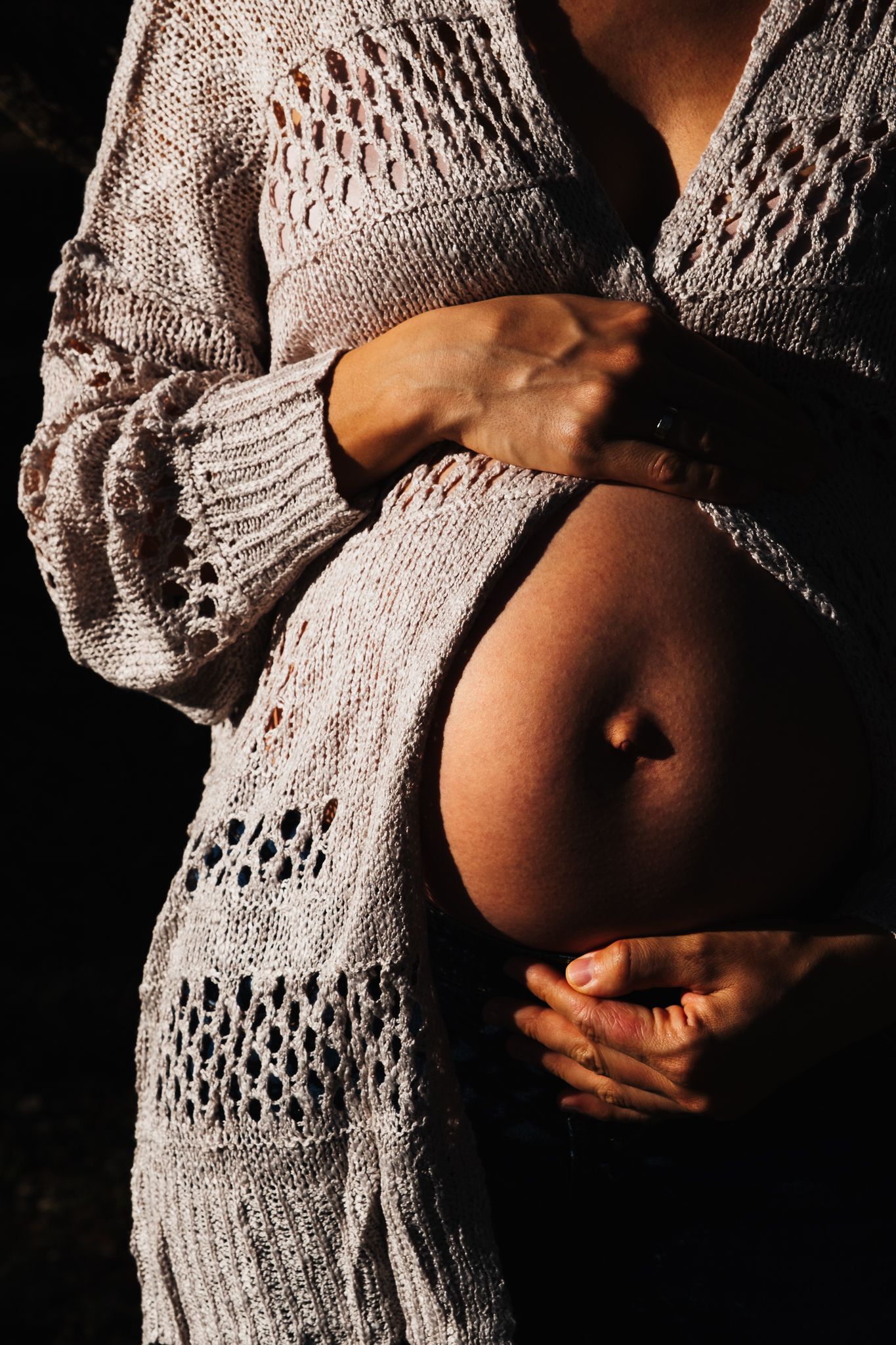 A close-up photograph of a pregnant belly being cradled in hands, wearing a cozy grey knit cardigan sweater.
