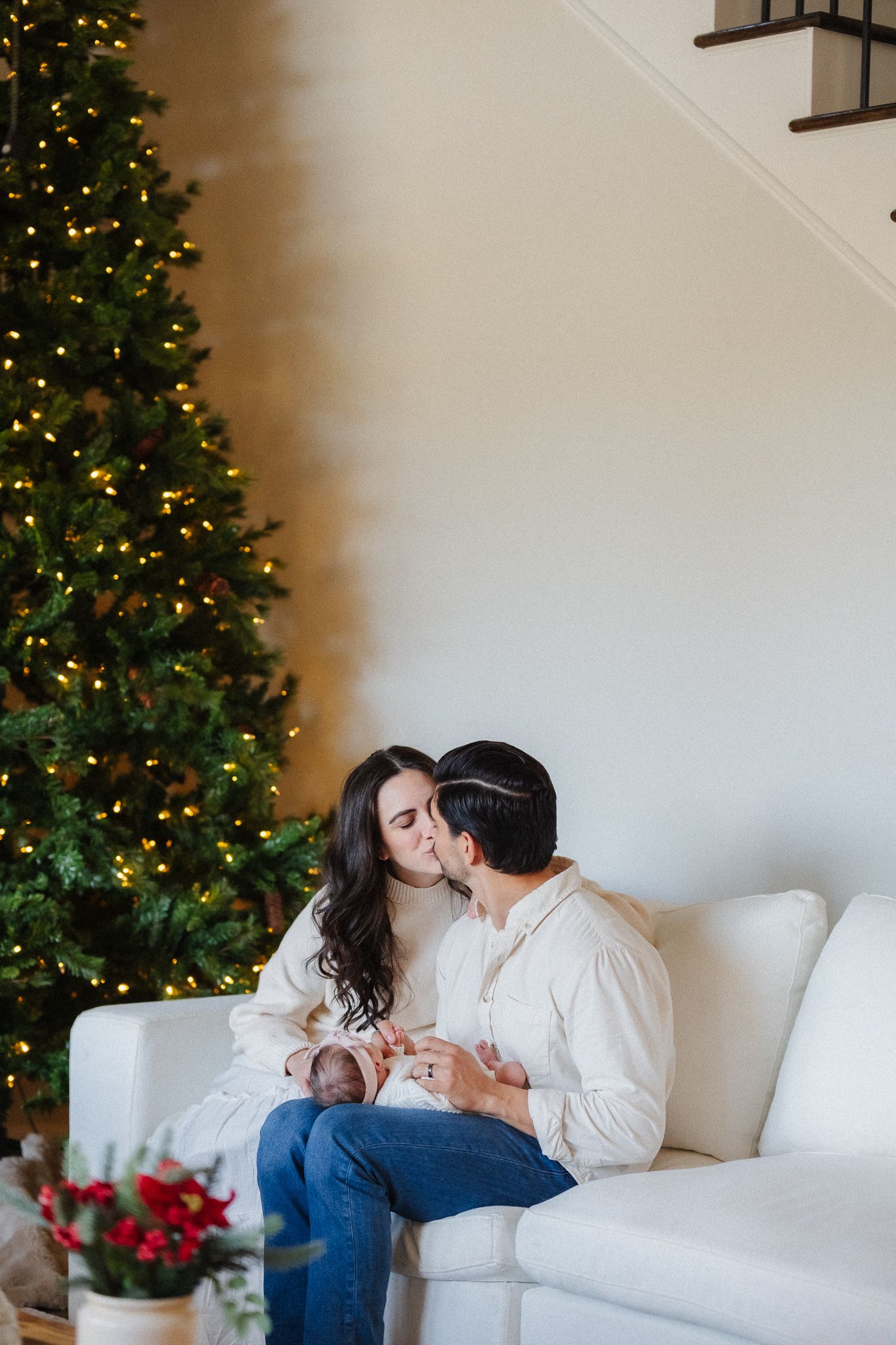 Couple sharing a kiss on white sofa with lit Christmas tree in background during holiday family photo session.