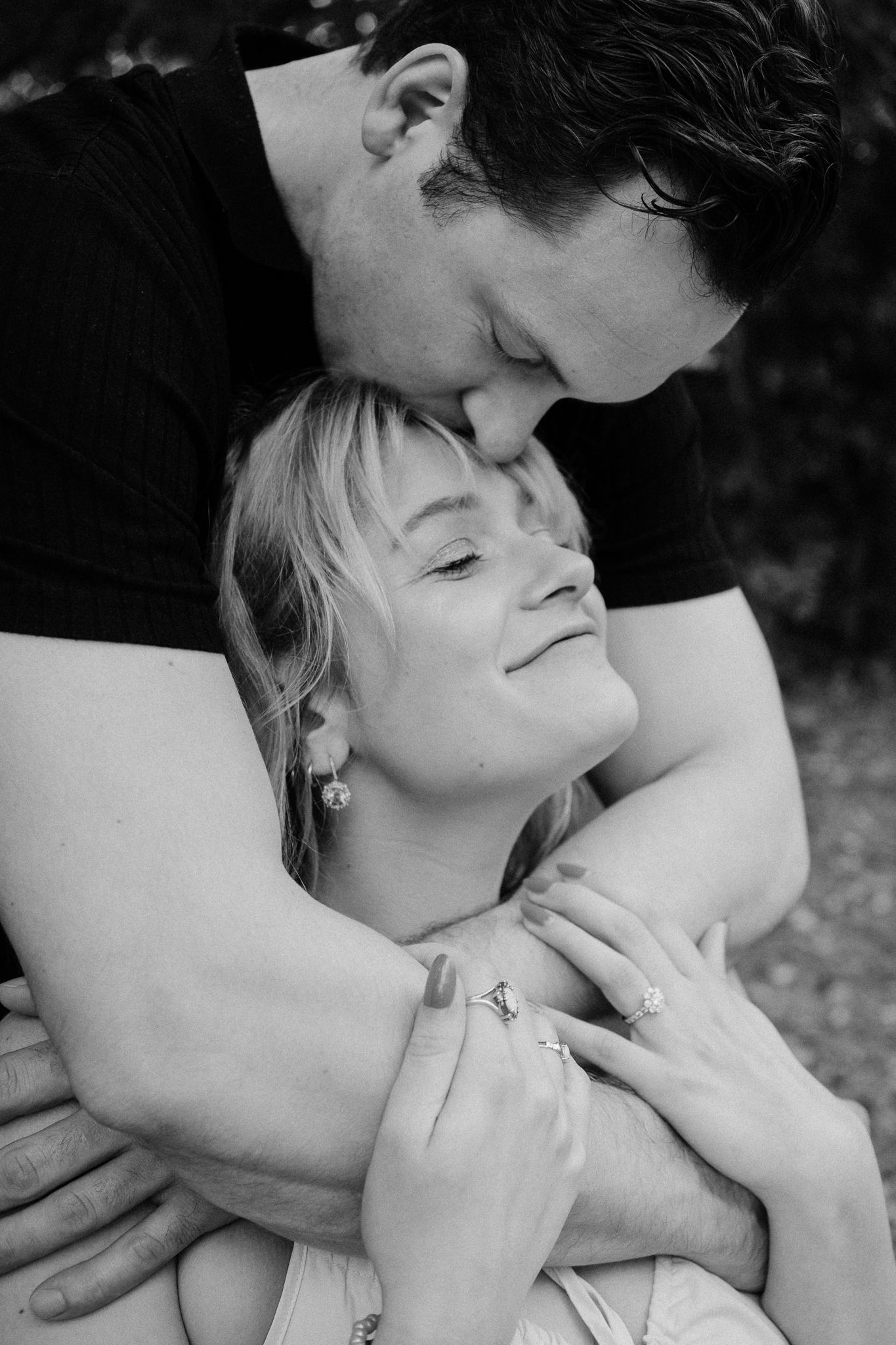 Black and white photo of a couple embracing, man kissing womans forehead as she smiles, showing engagement ring.