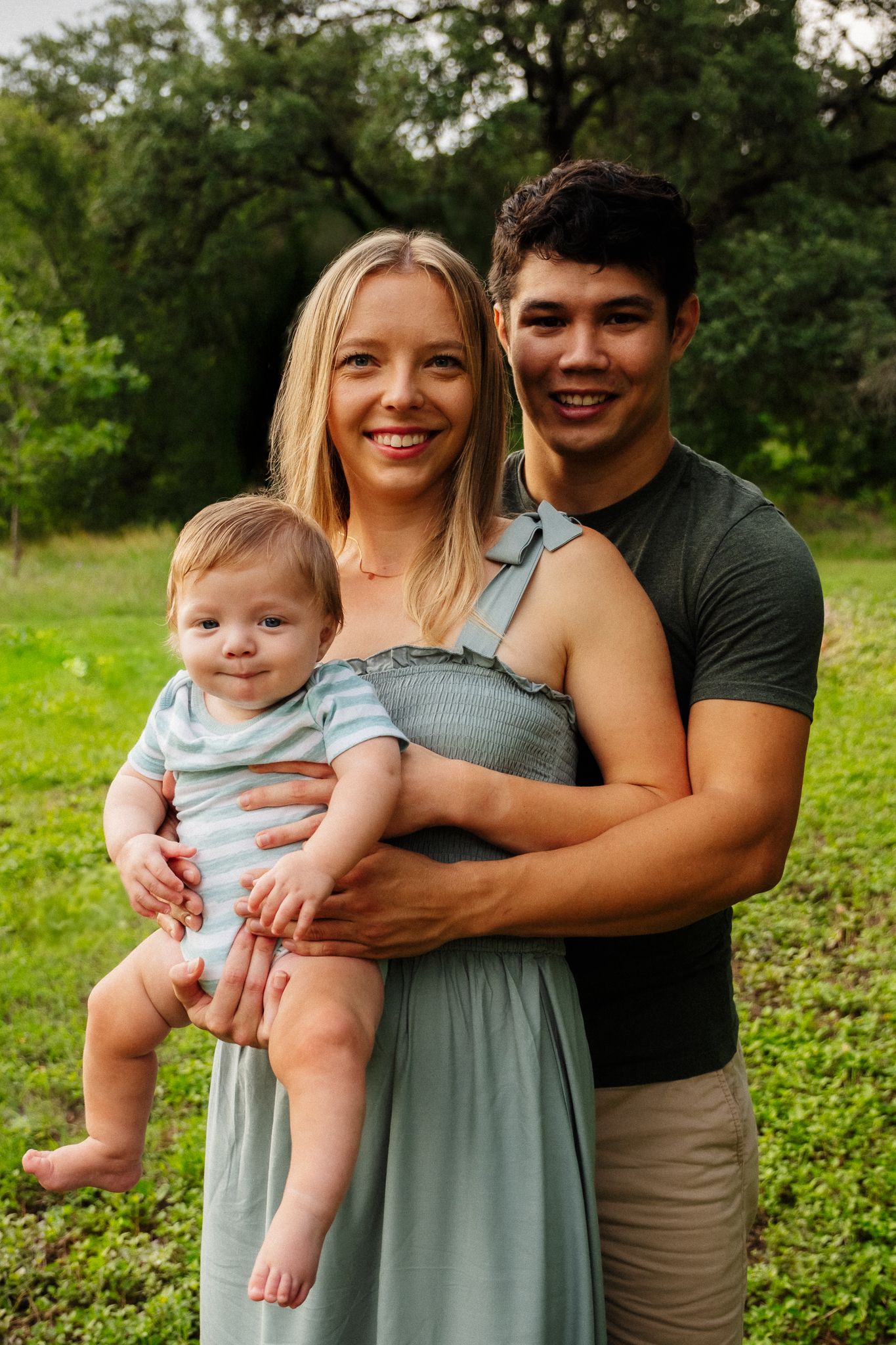 A family poses together in a lush green outdoor setting wearing casual clothes in matching earth tones.
