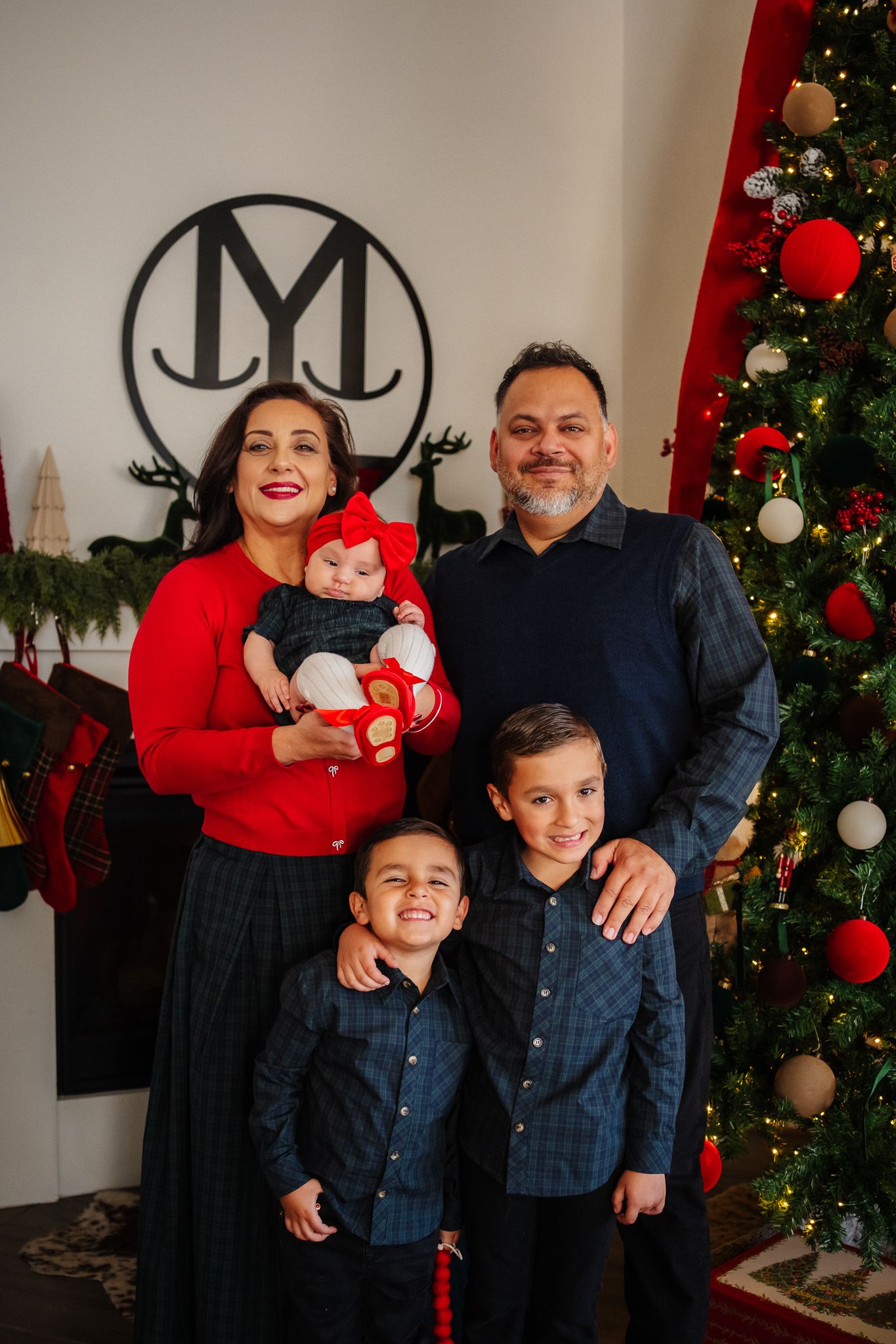 Family poses together in front of a decorated Christmas tree with red and white ornaments and a black JM monogram on the wall.
