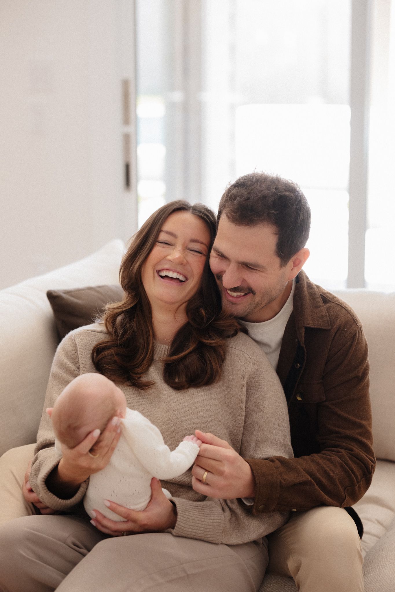 A couple shares joyful laughter while cuddling together on a light colored couch in a bright, airy room.