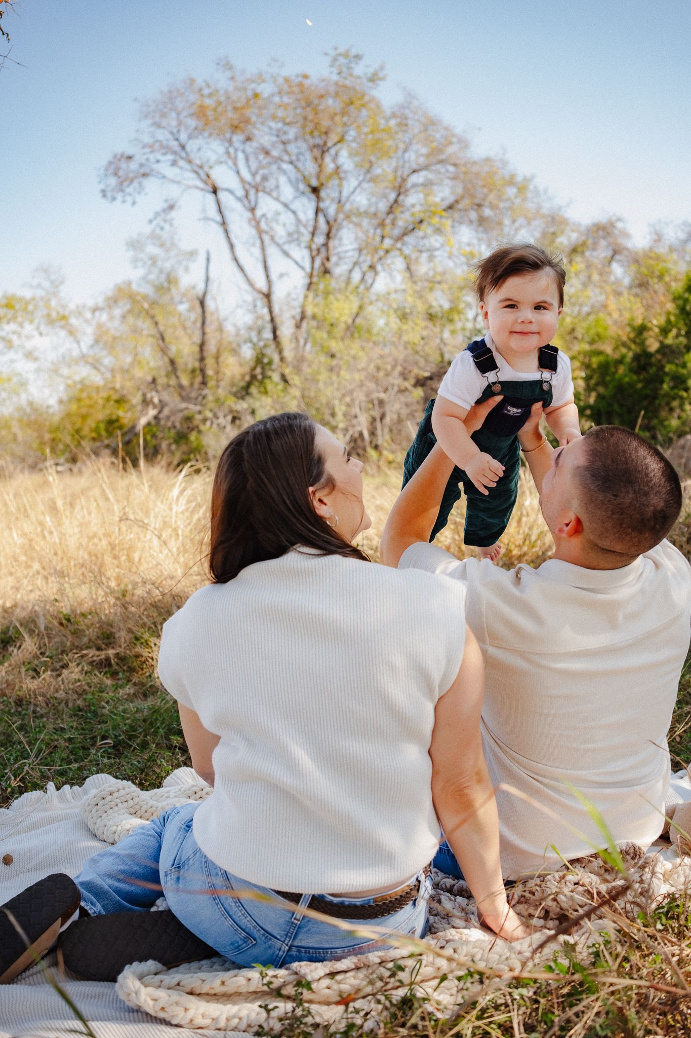 Family sitting together on a blanket in a sunny field taking photos and enjoying quality time outdoors.