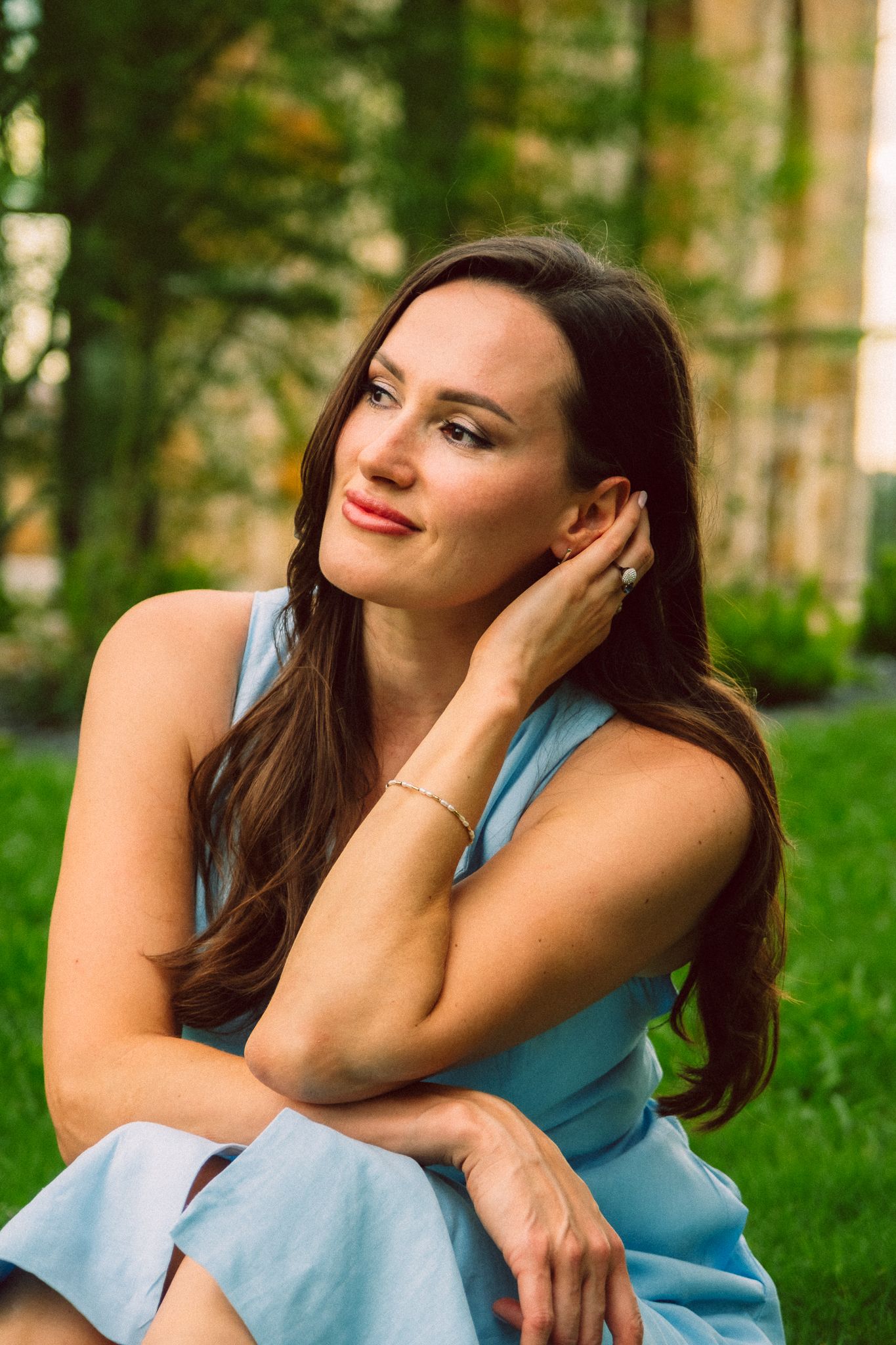 Woman in light blue dress poses thoughtfully on grass in a summer garden setting.