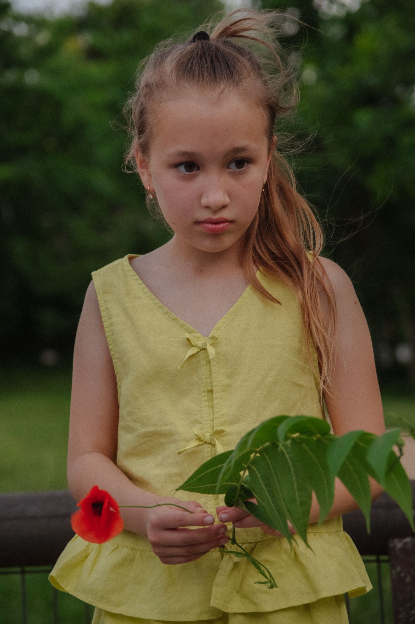 Child in yellow sundress holding foliage while standing outdoors near blooming red poppy flower on summer day.