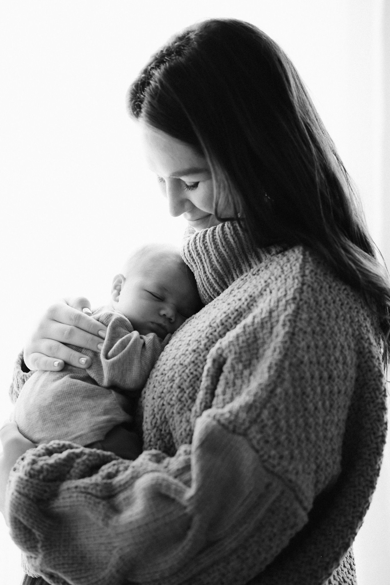 Black and white photo of a smiling mother in a knit sweater tenderly cradling her sleeping newborn baby.