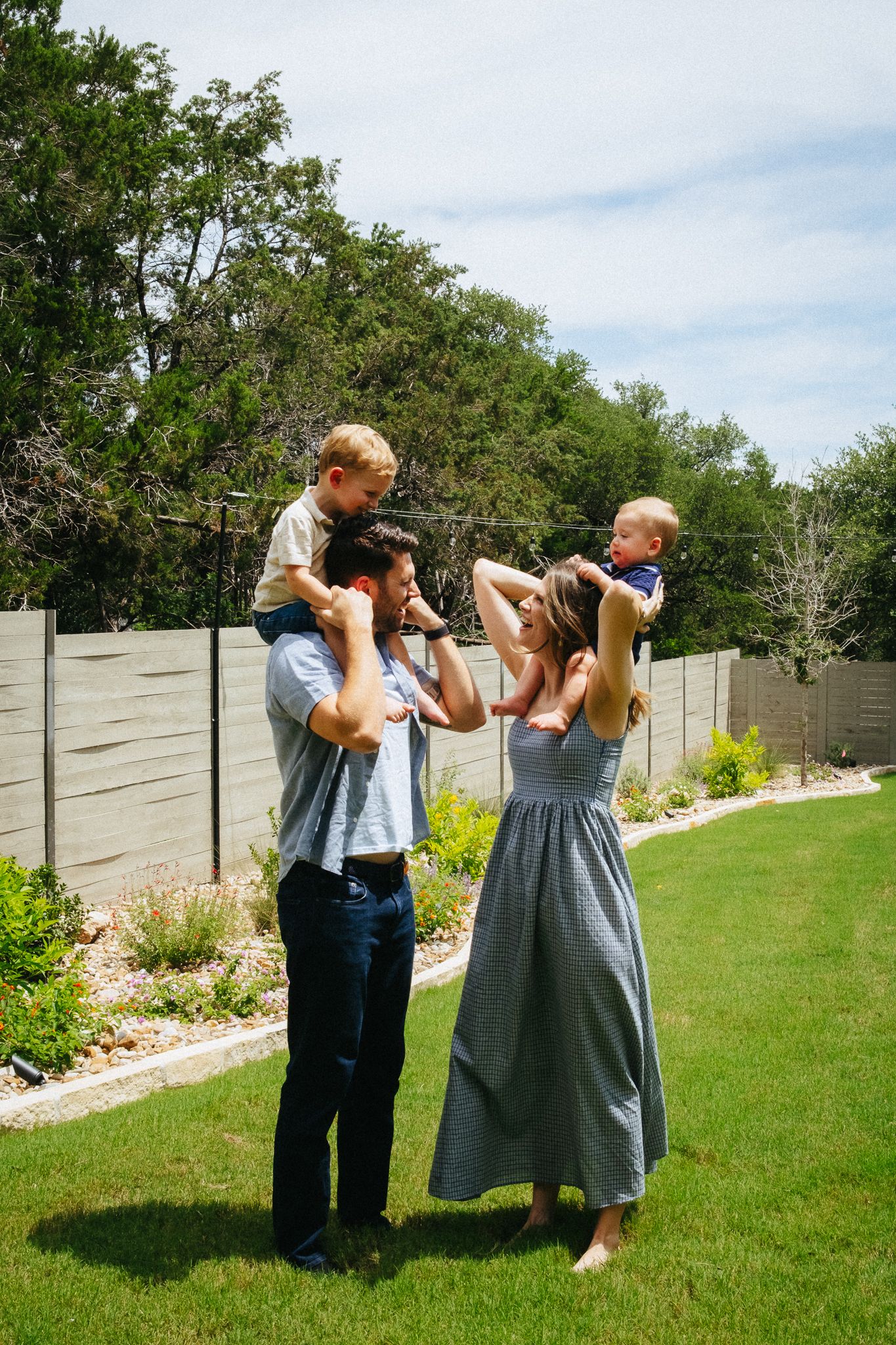 A happy family of four stands in a sunny backyard with both young children perched on their parents shoulders.