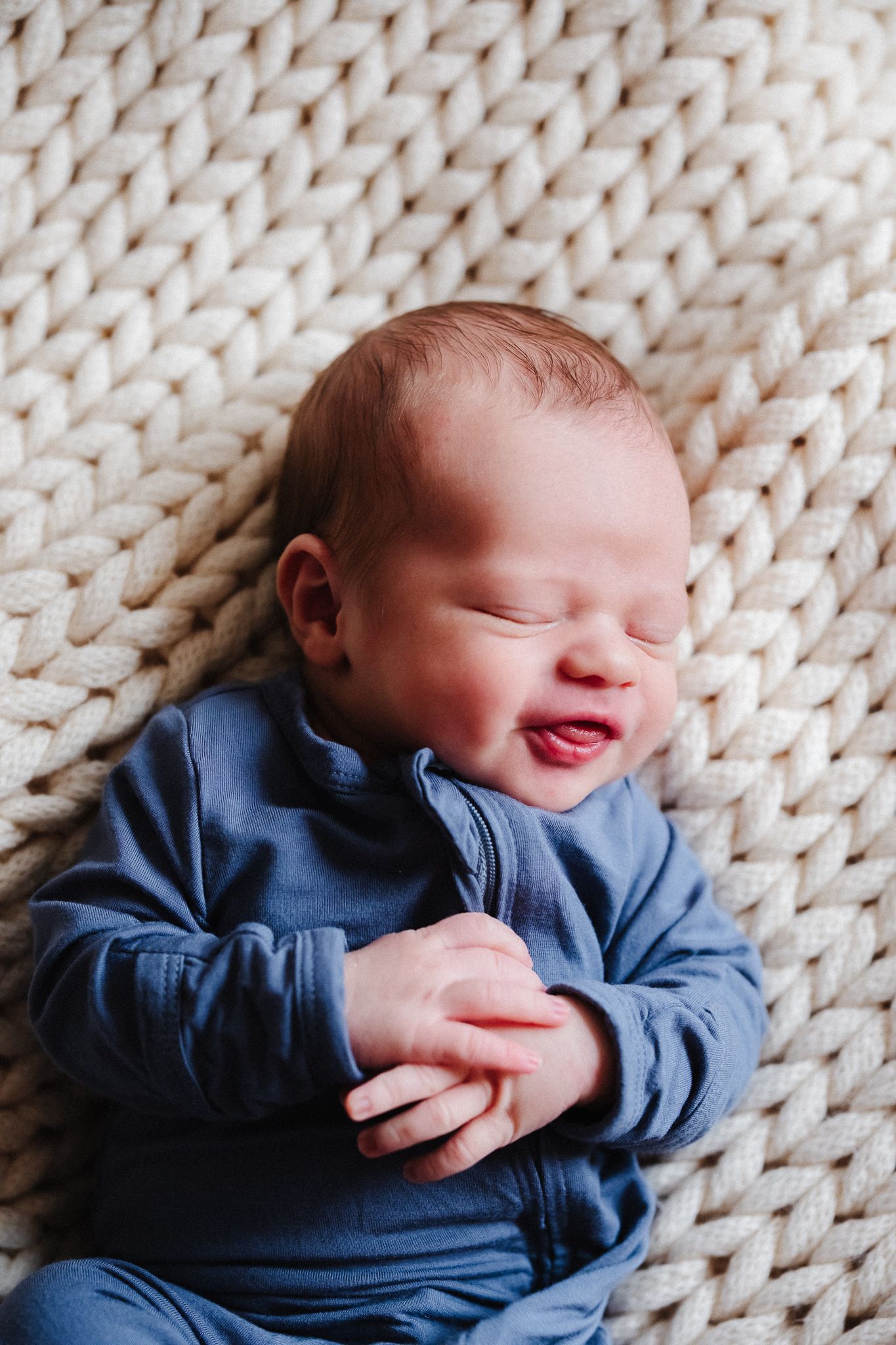 Smiling baby wearing blue sleepwear lies on chunky knit cream blanket during newborn photoshoot.