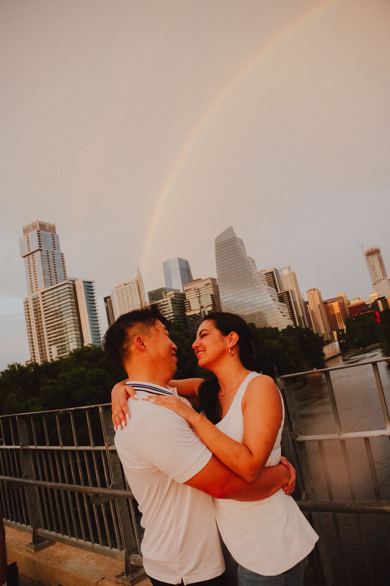 Couple embracing on a bridge with Austin skyline and rainbow in the background at golden hour.
