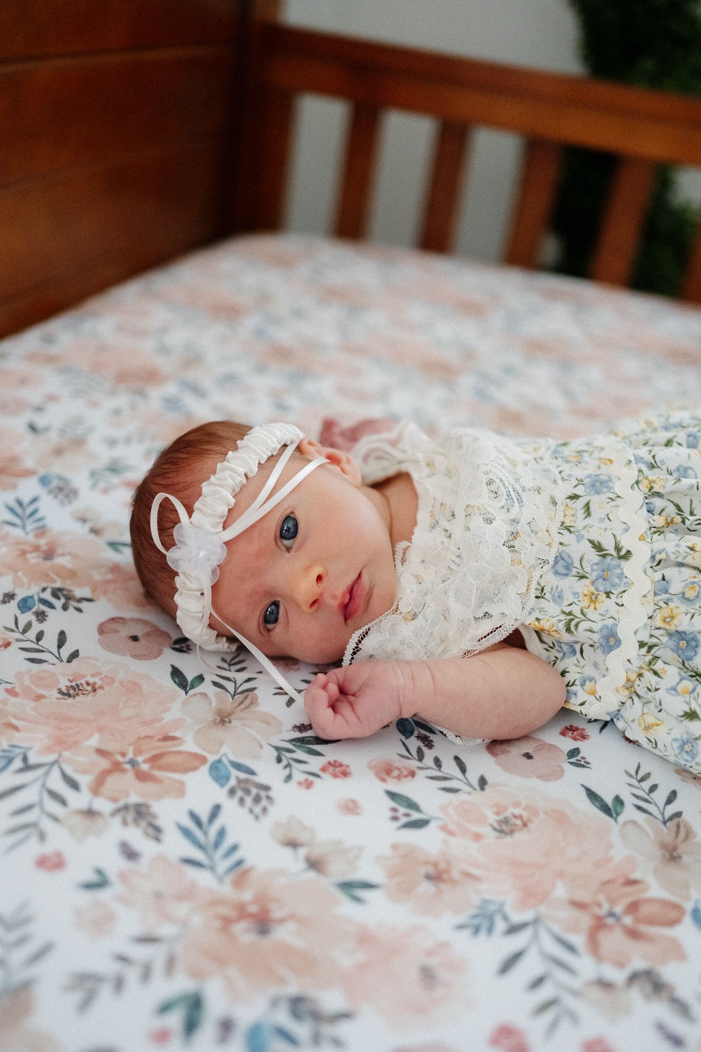 Newborn baby girl in floral wrap and lace headband lying on floral crib sheet, looking at camera.