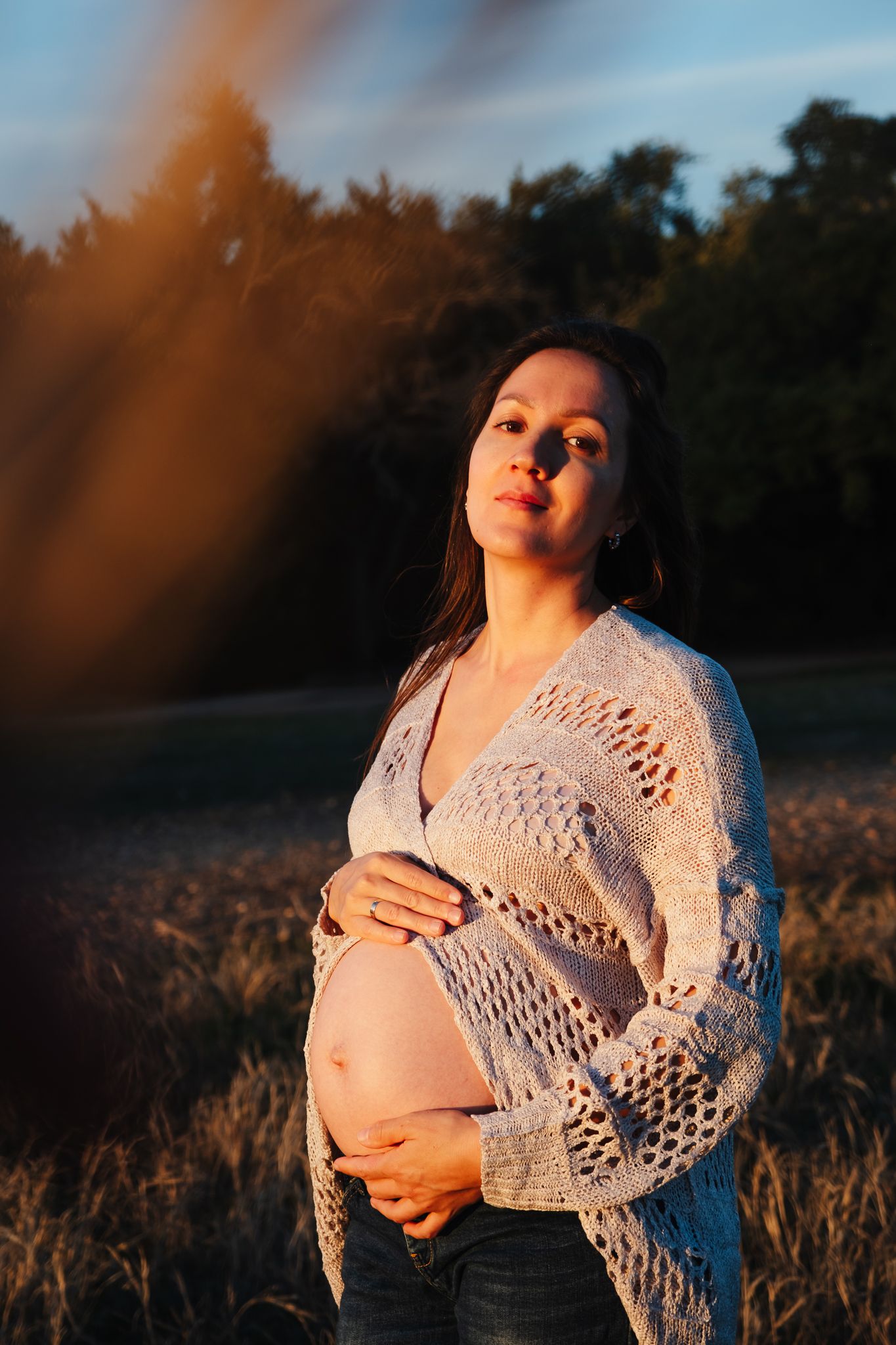 A pregnant woman stands in a wheat field at sunset wearing a white crocheted sweater and jeans.