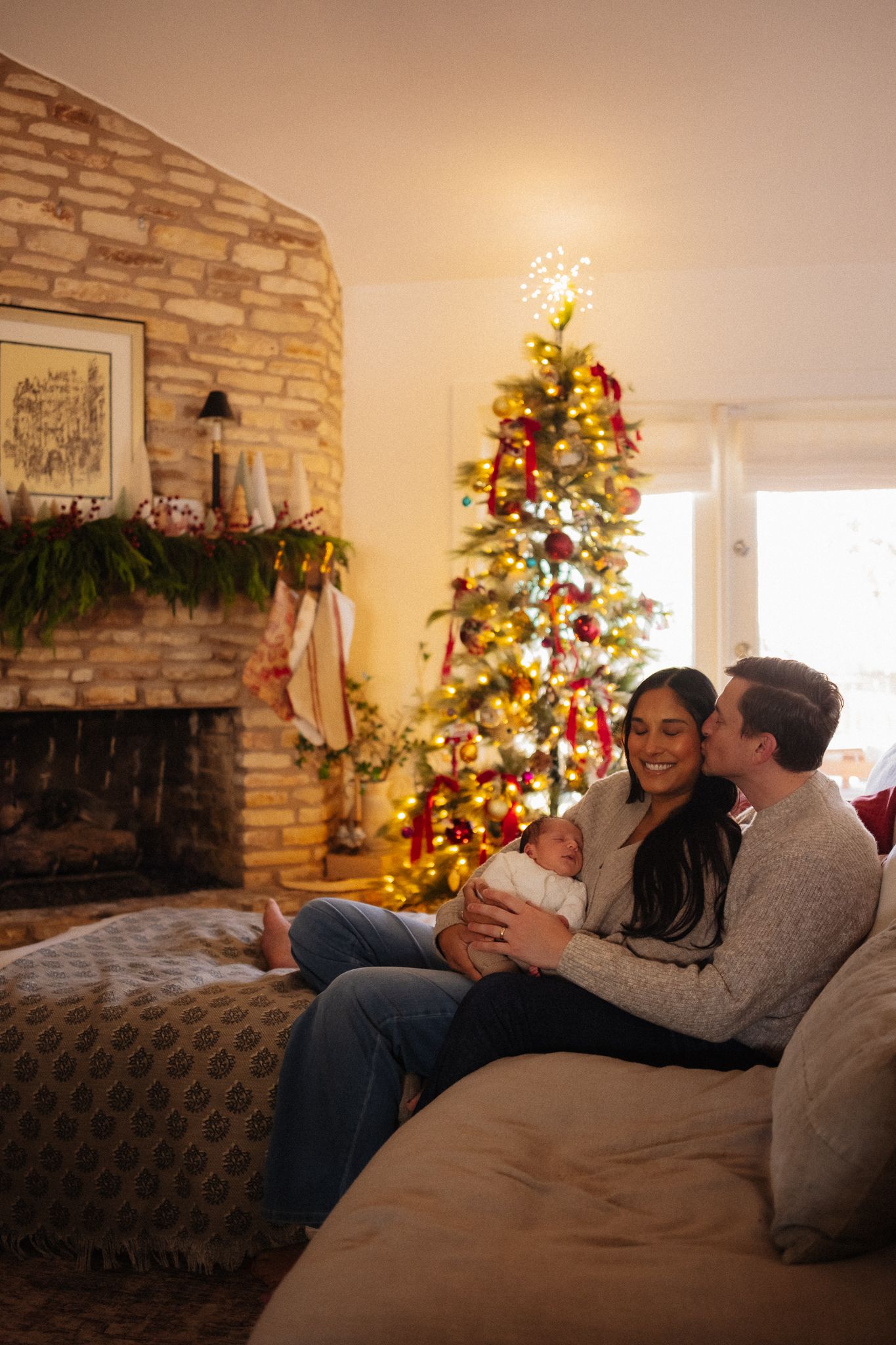 Happy couple sitting on couch holding newborn baby in festively decorated living room with Christmas tree.