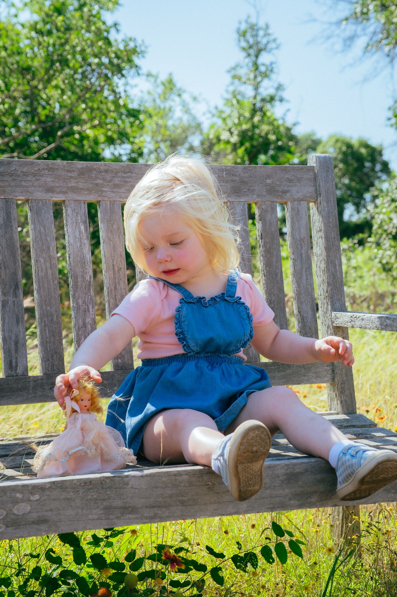 Small child sits and explores curiously on a wooden bench against sunny green summer landscape background.