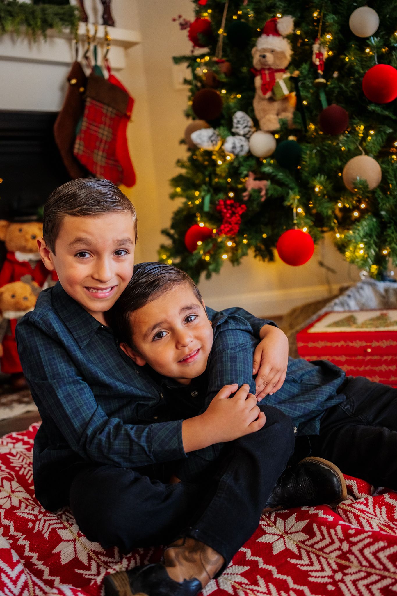Two children playfully pose on a red and white snowflake blanket in front of a decorated Christmas tree.