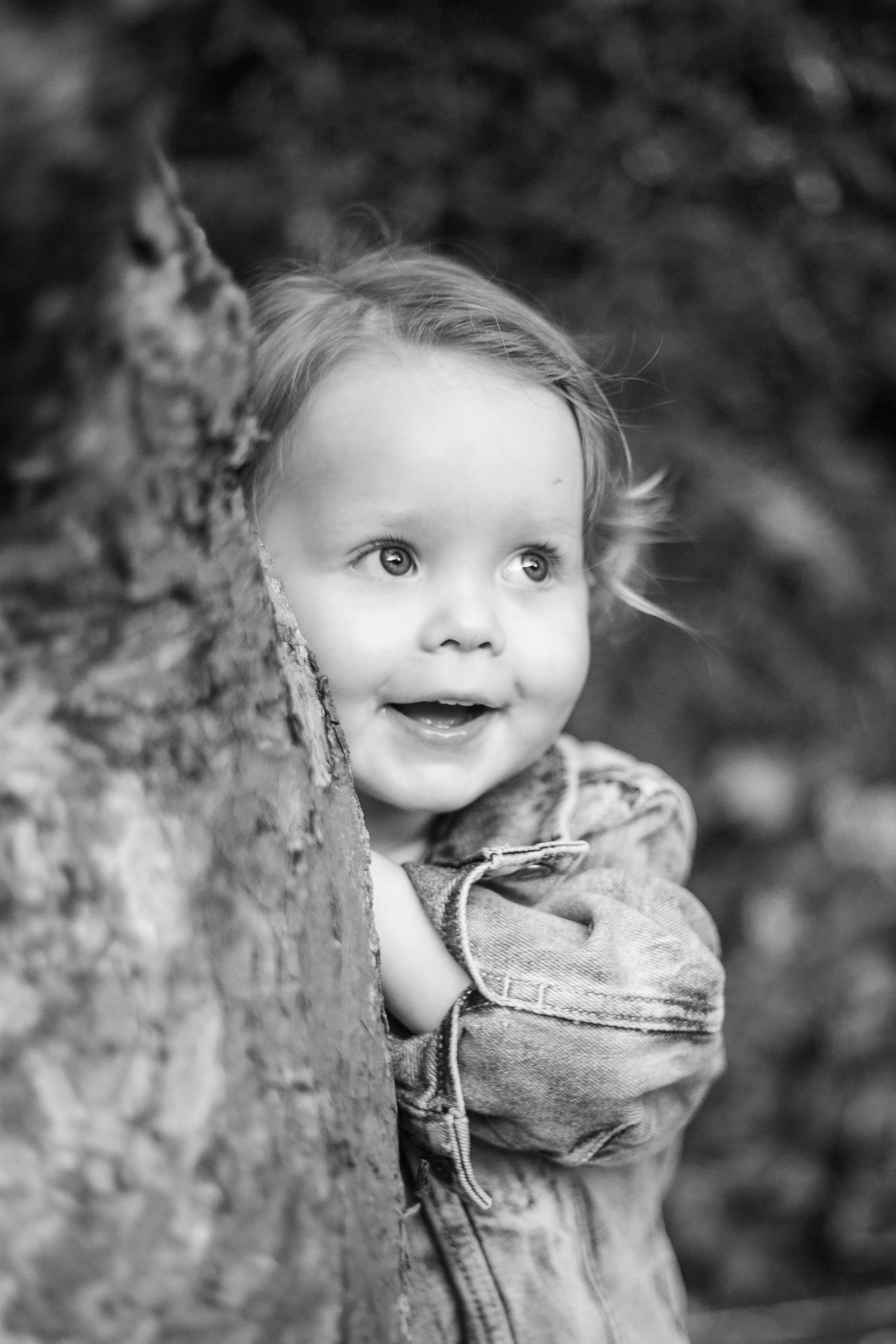 Black and white portrait of a joyful young child peeking around a tree trunk.