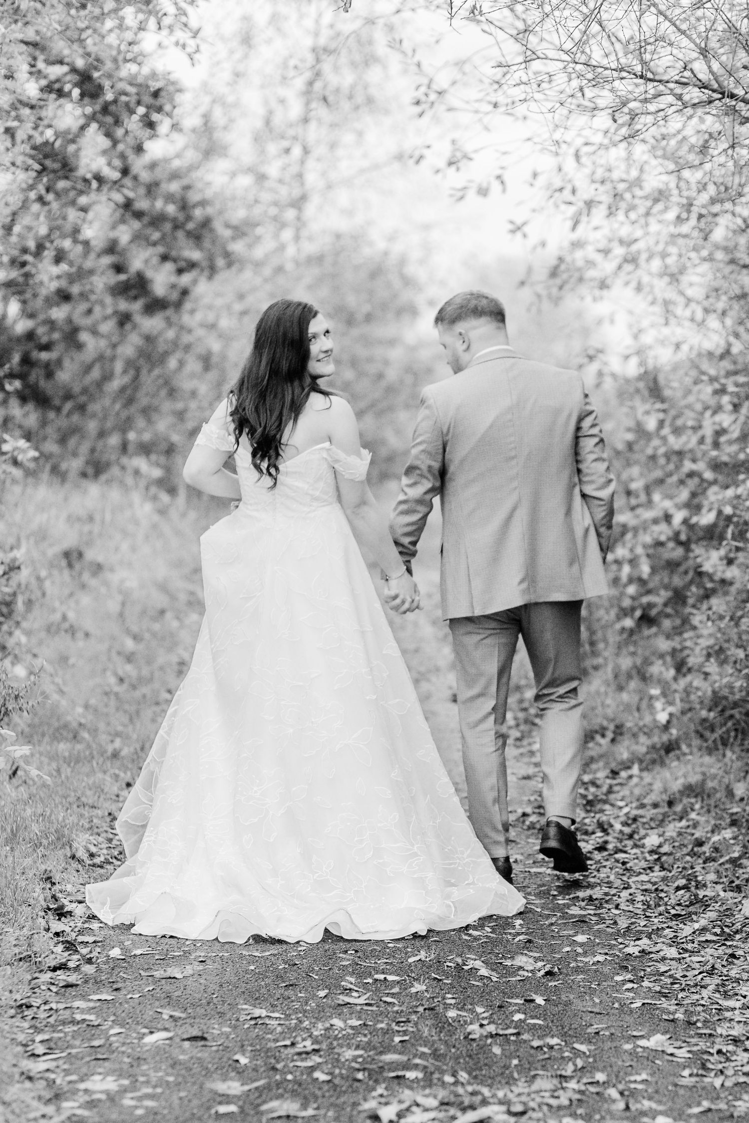 Black and white photo of a couple walking down a nature path on their wedding day, her white dress trailing behind them.