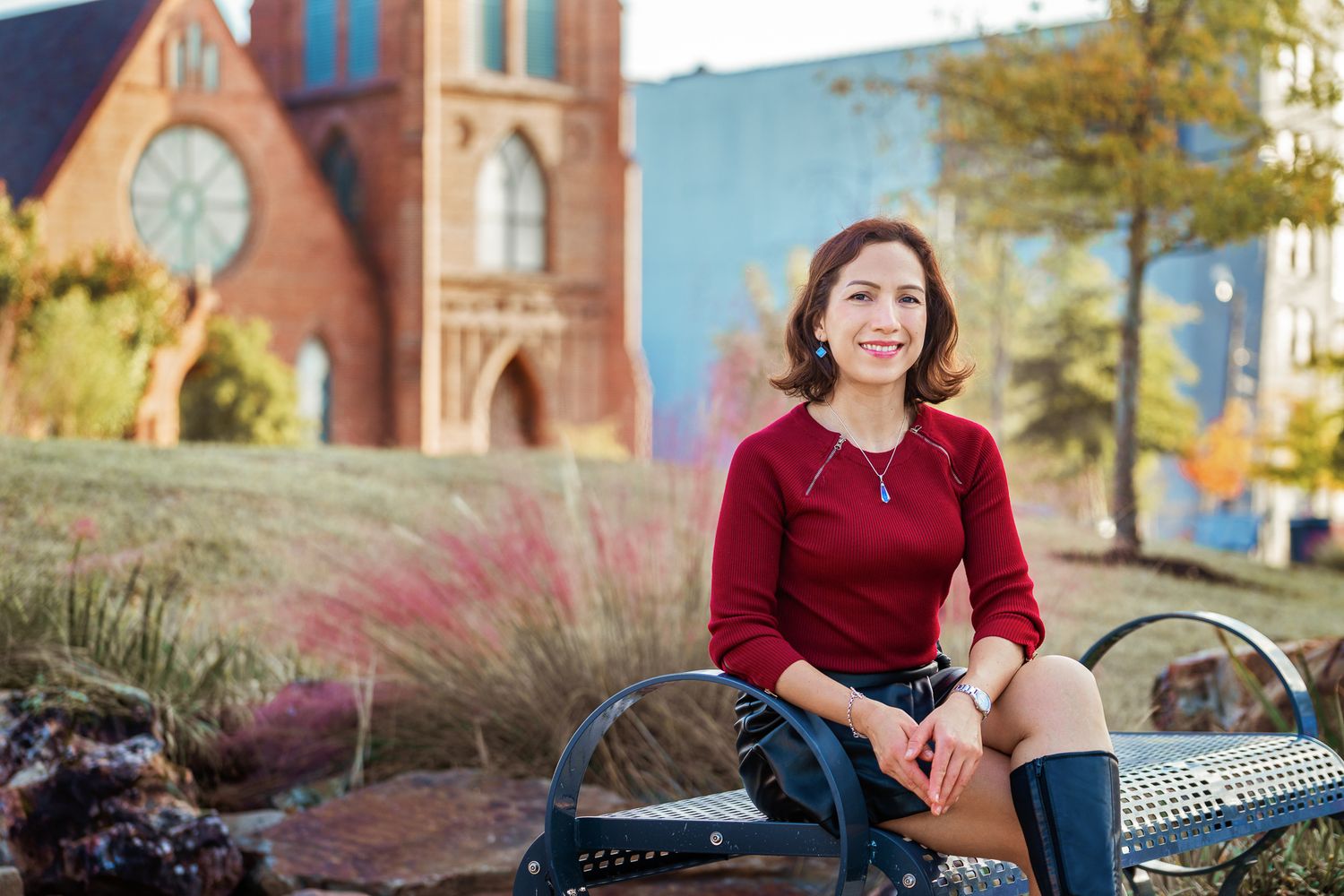 A person in a red sweater sits on a bench outside a brick church with ornate architecture and decorative grasses nearby.