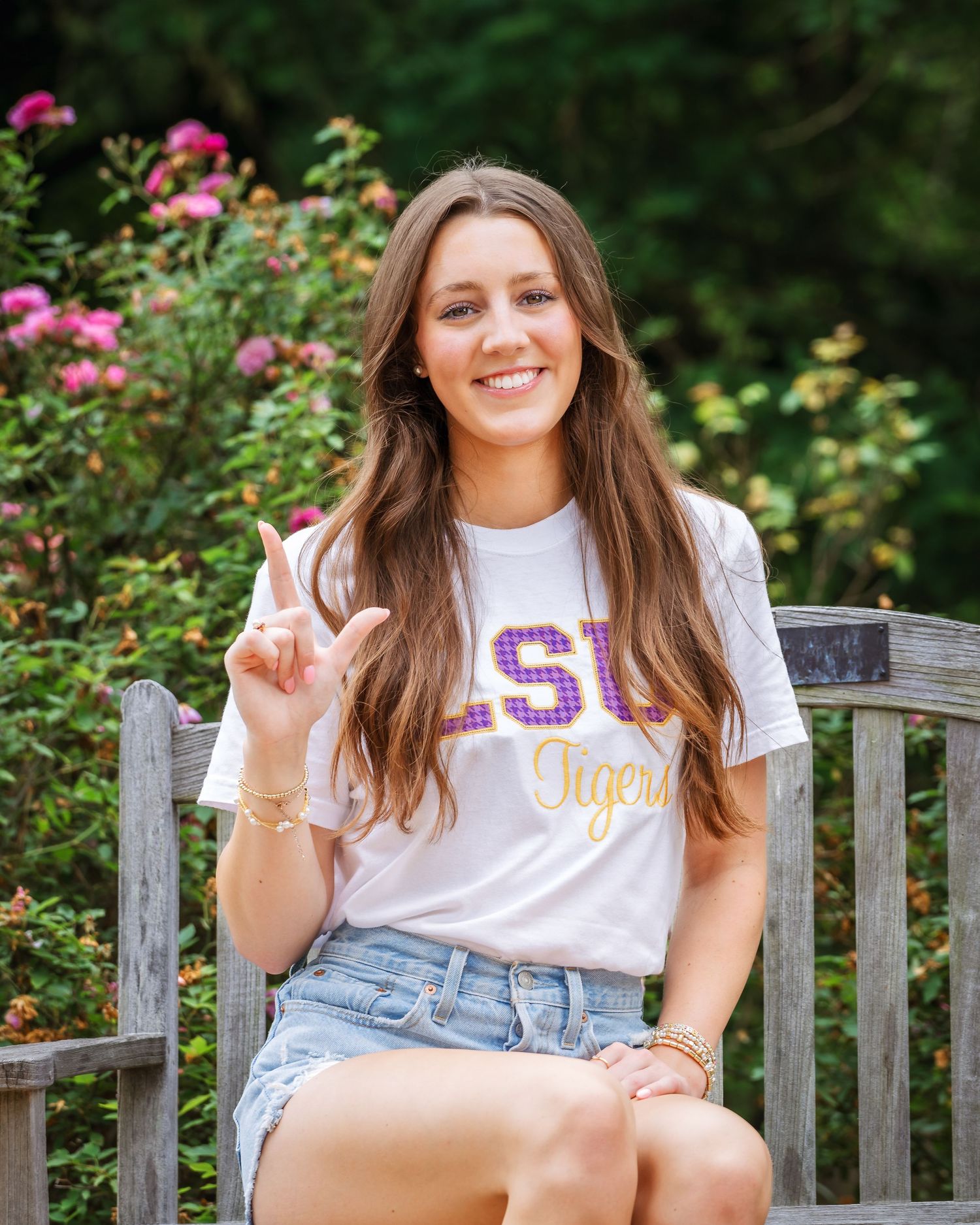 Student sits on wooden bench wearing LSU Tigers t-shirt and denim shorts with pink rose bushes in garden background.