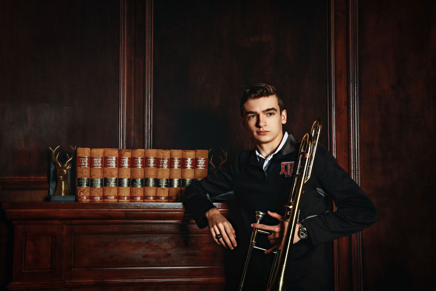 A person poses in a dark wood-paneled room wearing formal attire with leather seating and bookshelves in the background.