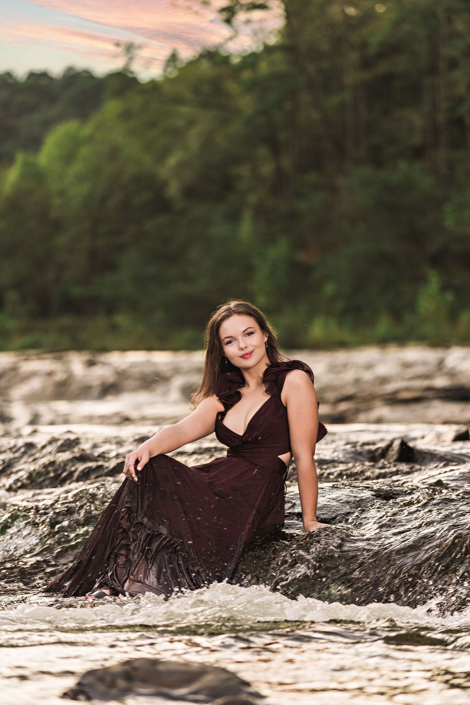 A woman in a burgundy dress poses gracefully on rocky riverbank during sunset.