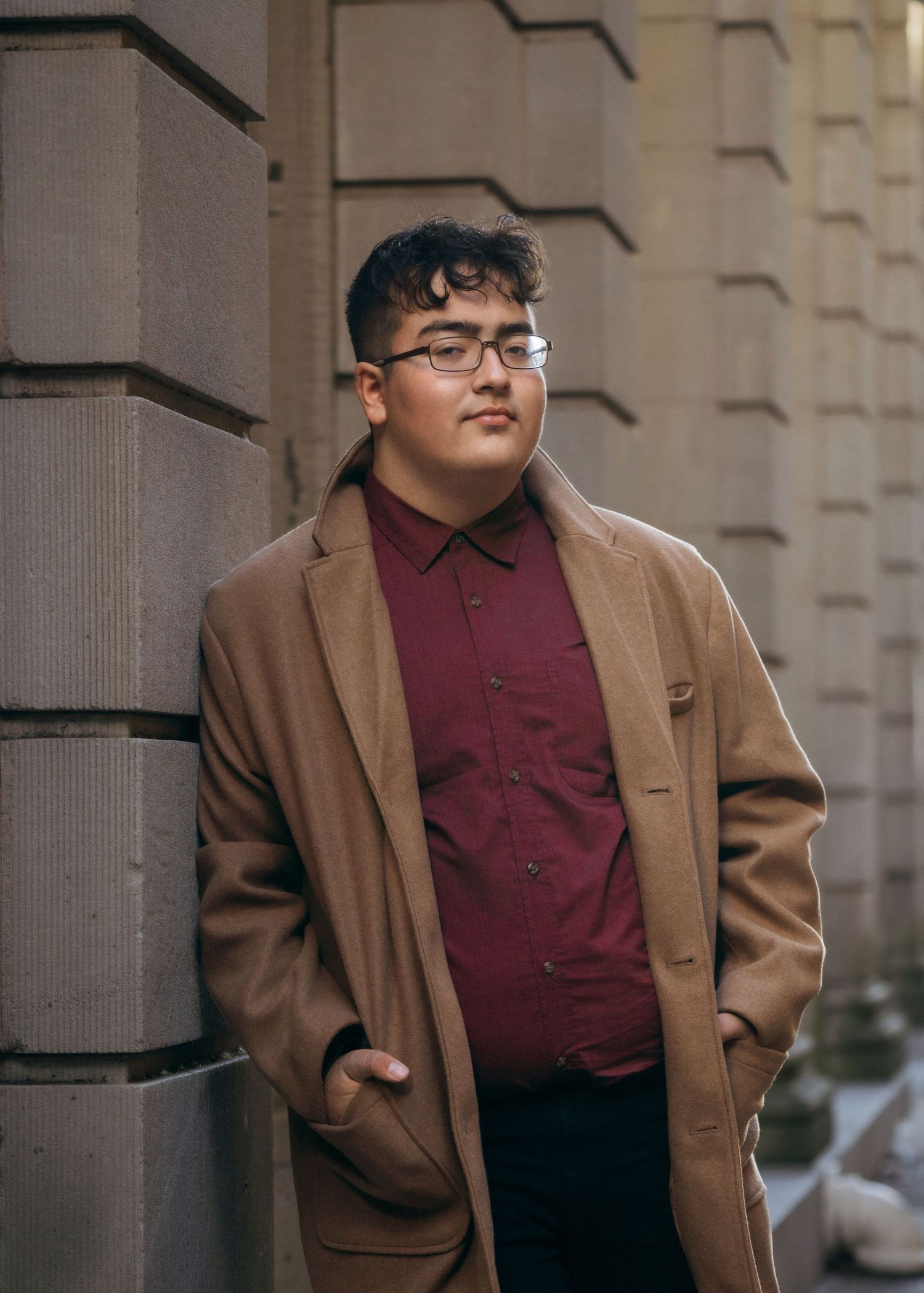 Young person in glasses wearing a burgundy button-down shirt and tan coat poses against a stone building exterior.
