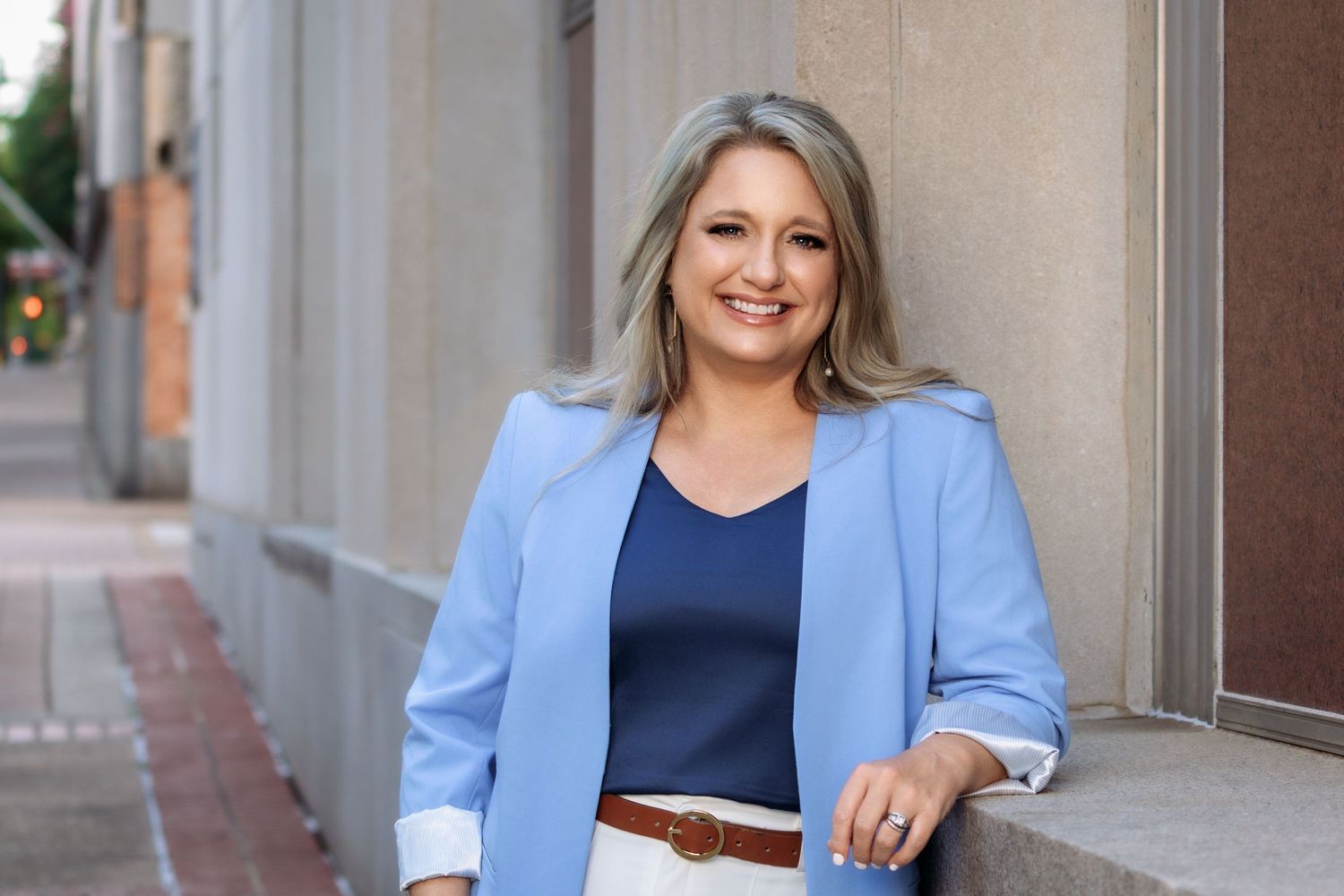 Professional in light blue blazer and navy top smiles while standing against urban building exterior.