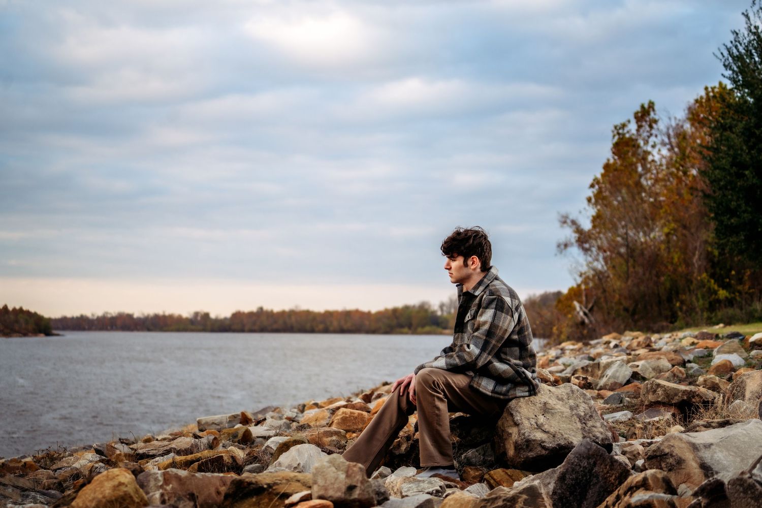 Someone sits peacefully on rocky shoreline by calm lake during autumn sunset wearing leather jacket.
