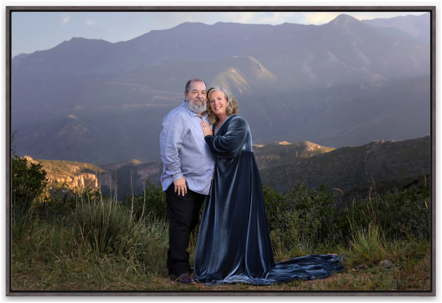 A couple in formal attire embraces outdoors against a backdrop of scenic mountain peaks at sunset.